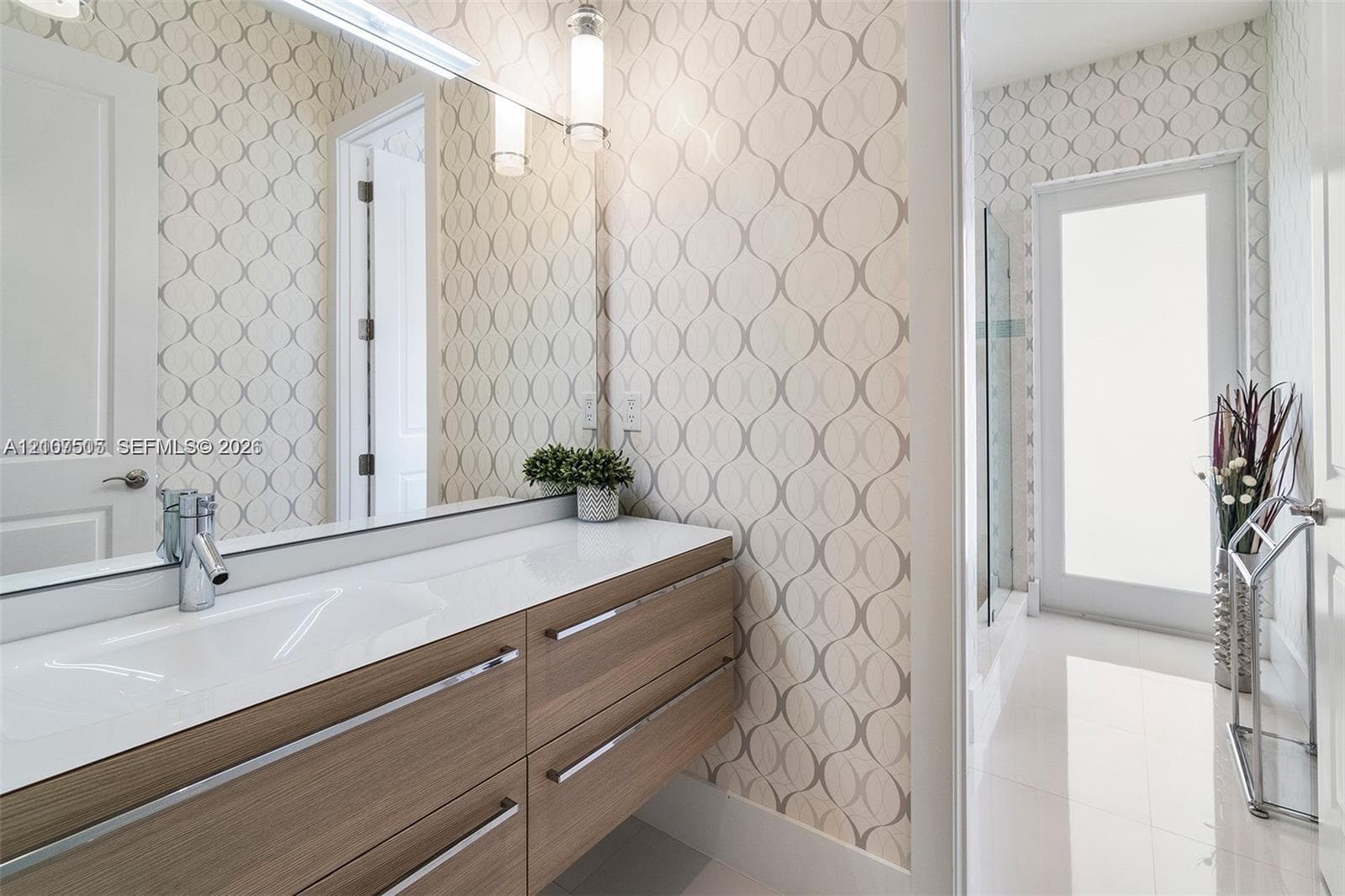 Floating wood-finish vanity with an integrated basin, polished metal hardware, expansive wall-mounted mirror, and glass-enclosed shower featuring patterned wall coverings and large-format tile flooring