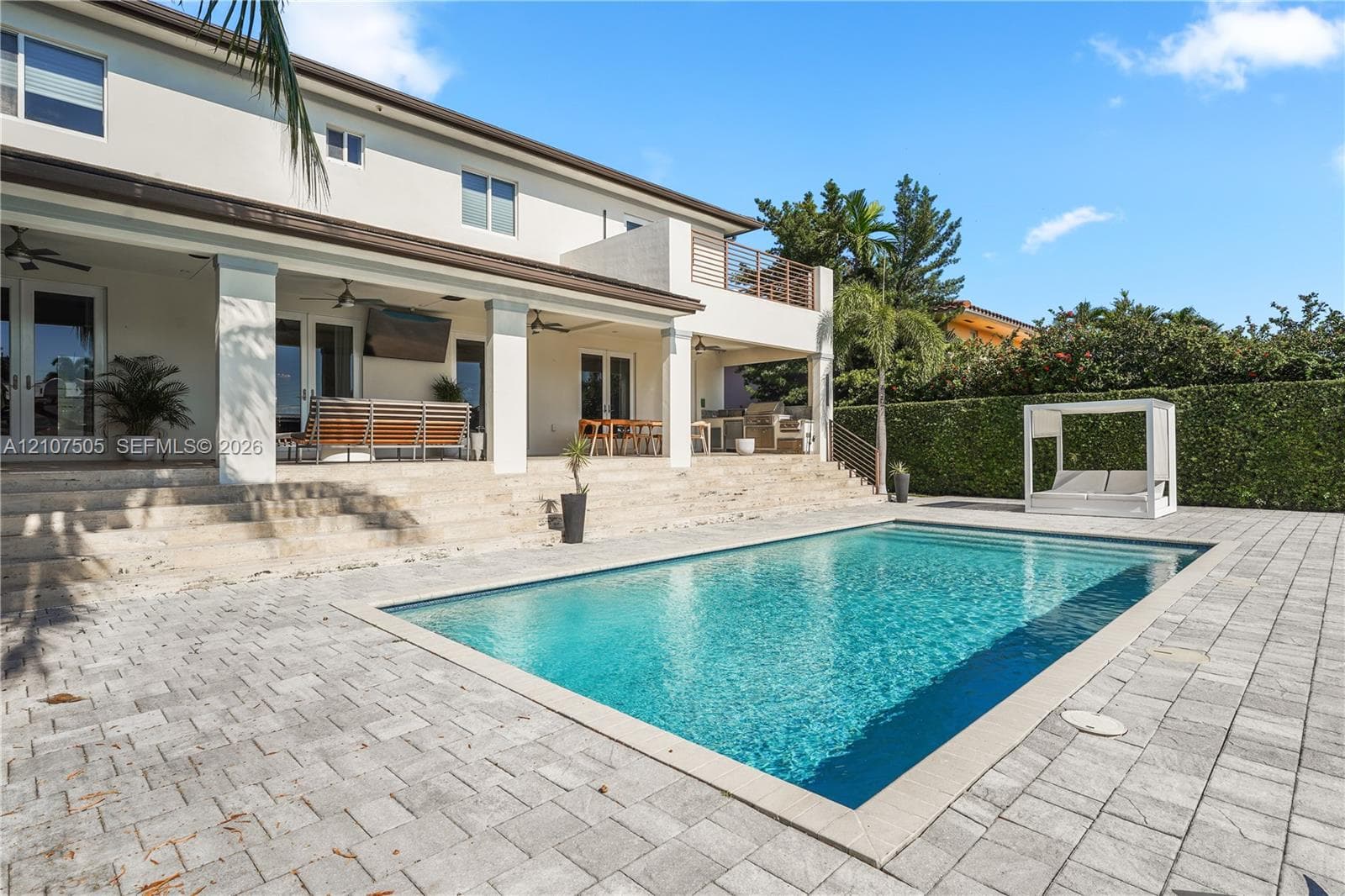 Covered rear patio with stone-finish steps, rectangular swimming pool, concrete paver decking, and upper-level balcony with horizontal railing