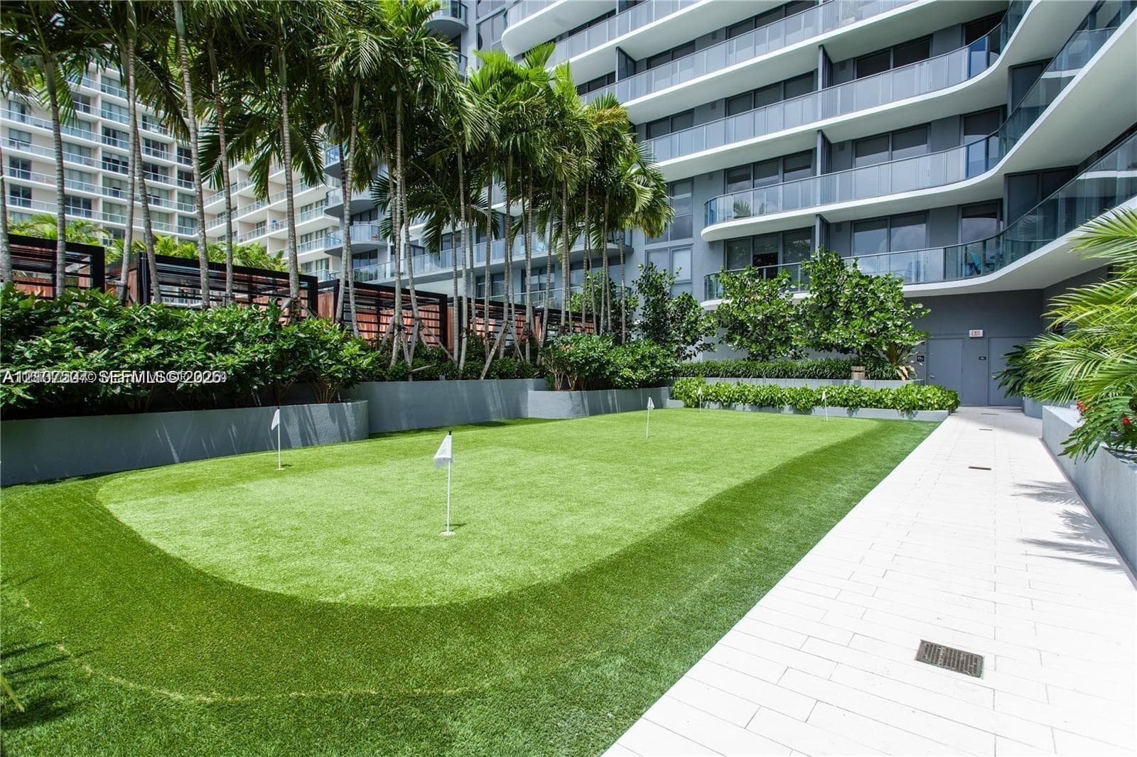 Putting green featuring synthetic turf, multiple flag pins, concrete paver walkways, mature palm trees, and perimeter privacy fencing