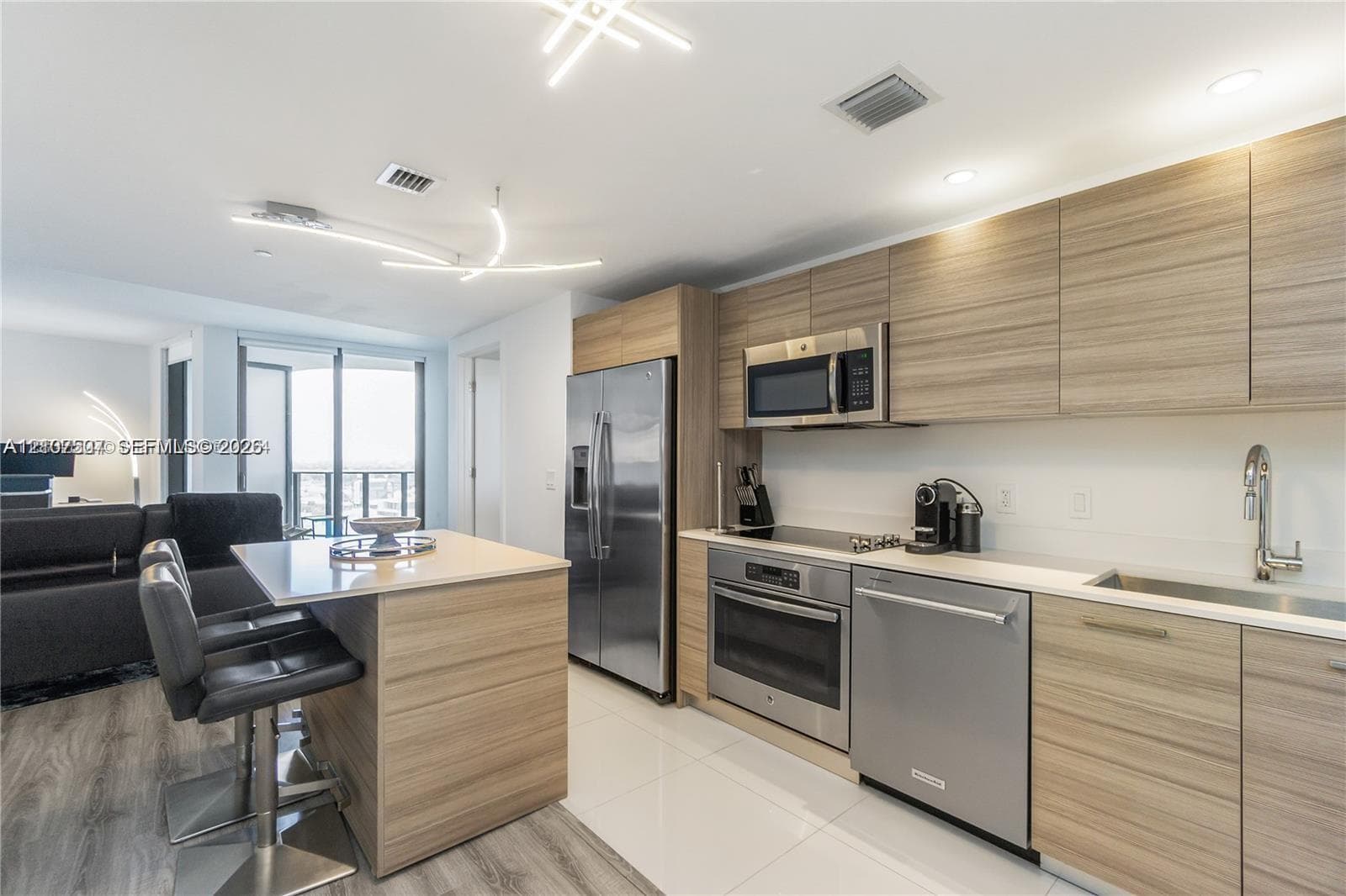 Modern kitchen featuring horizontal-grain wood-finish cabinetry, stainless steel appliances, quartz countertops, a matching island with integrated seating, and geometric LED lighting fixtures