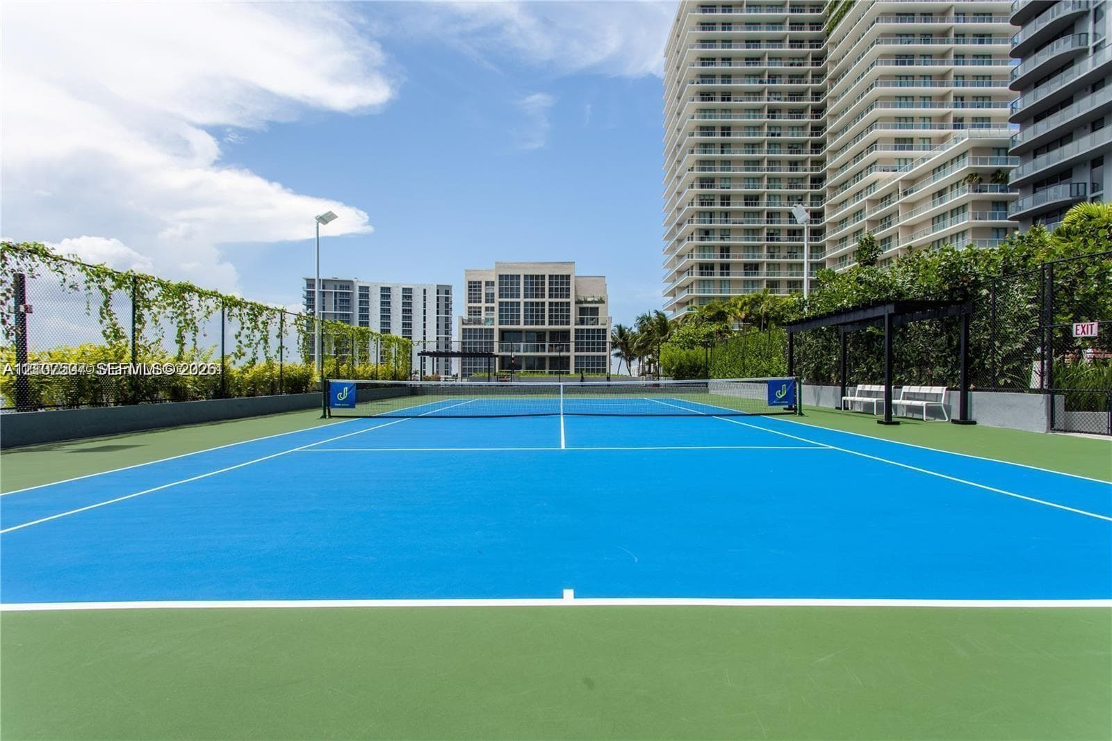 Tennis court featuring a hard surface, perimeter trellis fencing with climbing vegetation, and a covered bench area