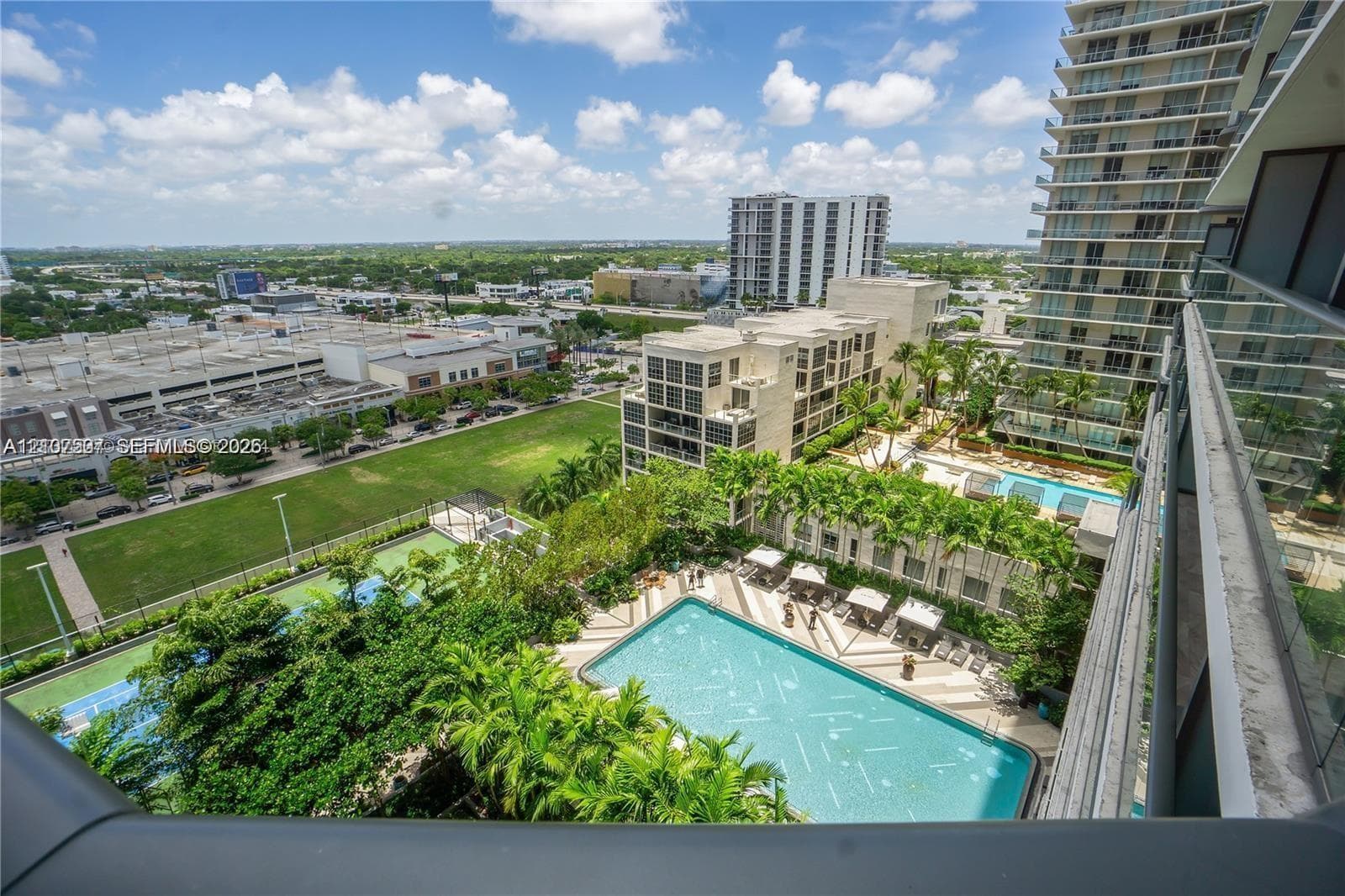 High-rise balcony featuring a glass panel railing, panoramic urban horizon, rectangular swimming pool, manicured palm trees, and hardscaped deck area