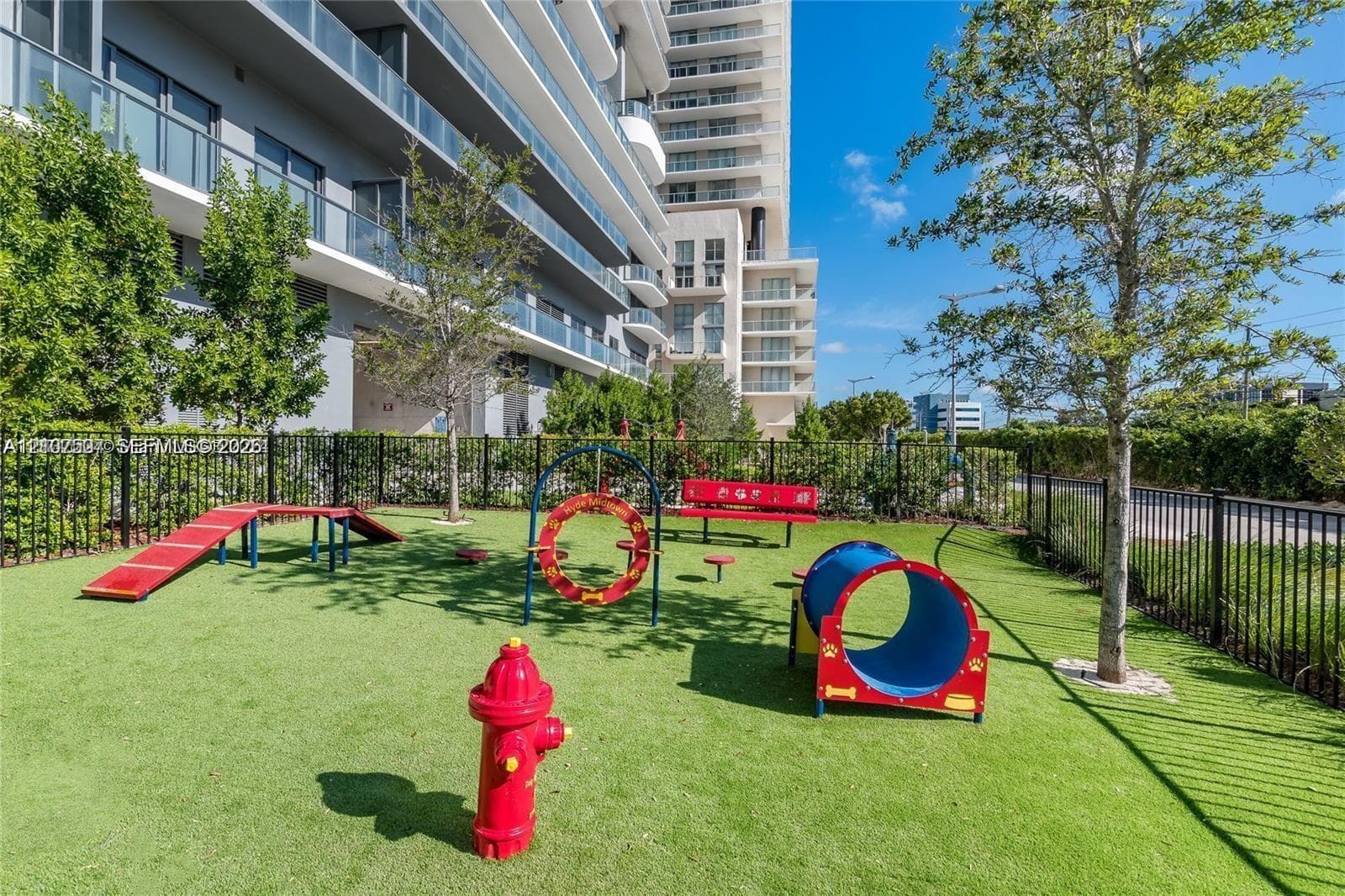 Outdoor pet recreation area featuring synthetic turf, metal perimeter fencing, and mature trees