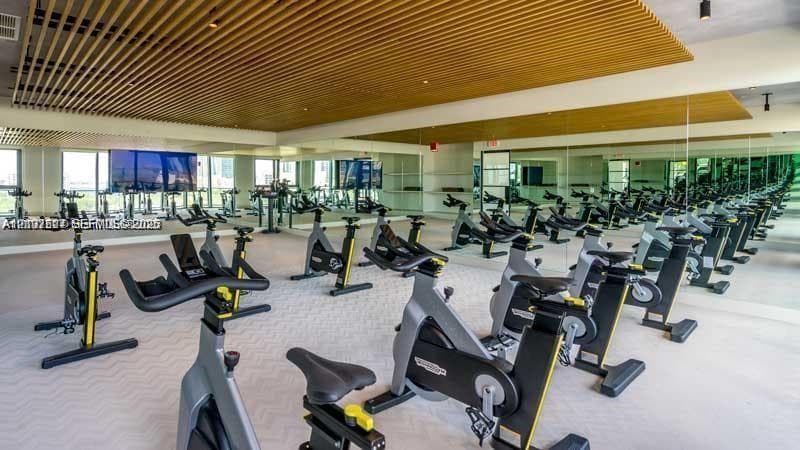 Fitness center featuring wood-slat ceiling detailing, floor-to-ceiling mirrors, large-format windows, and neutral patterned flooring