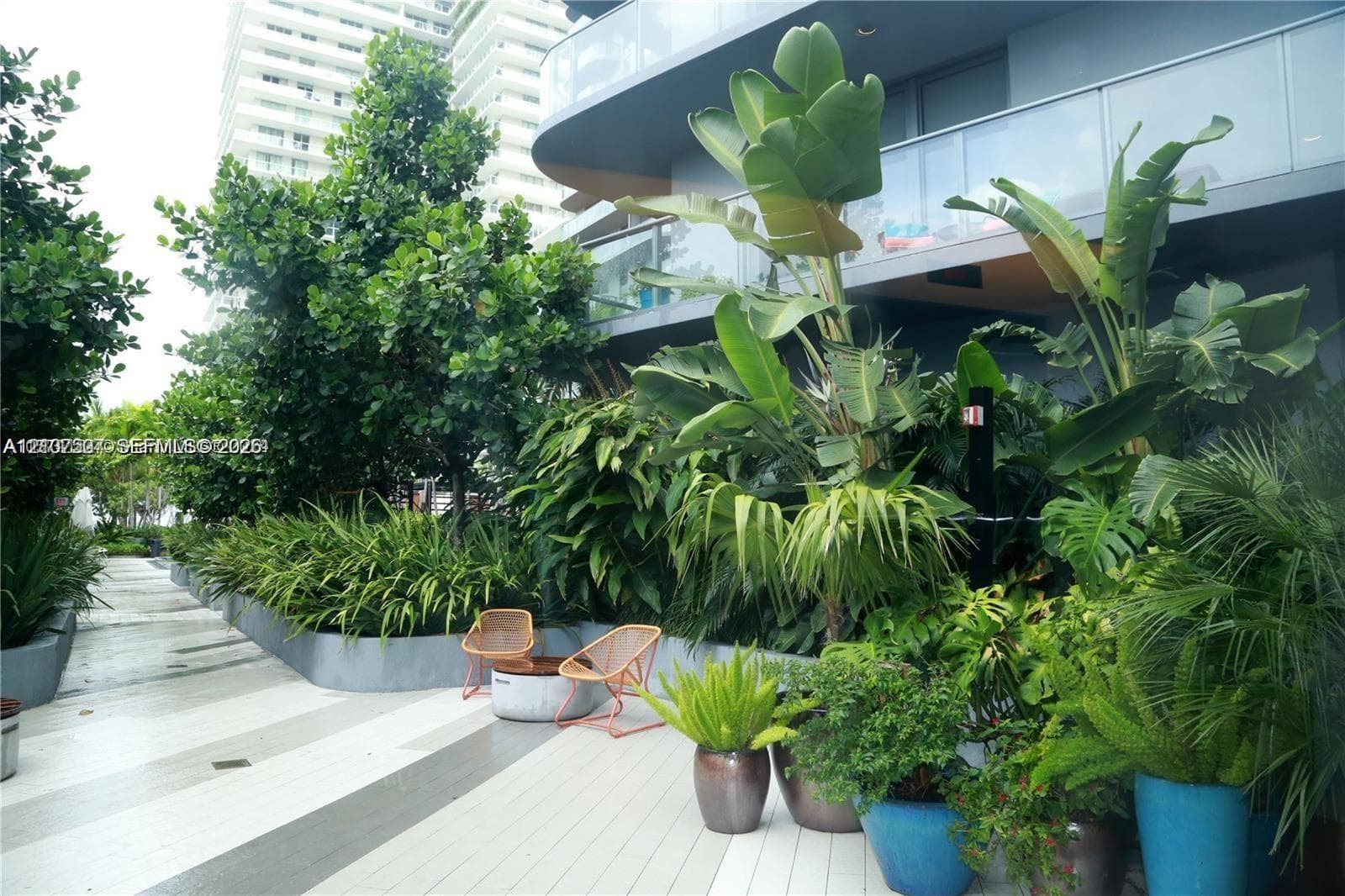 Private terrace featuring large-format tile flooring, perimeter planters with mature tropical landscaping, and a glass-paneled railing