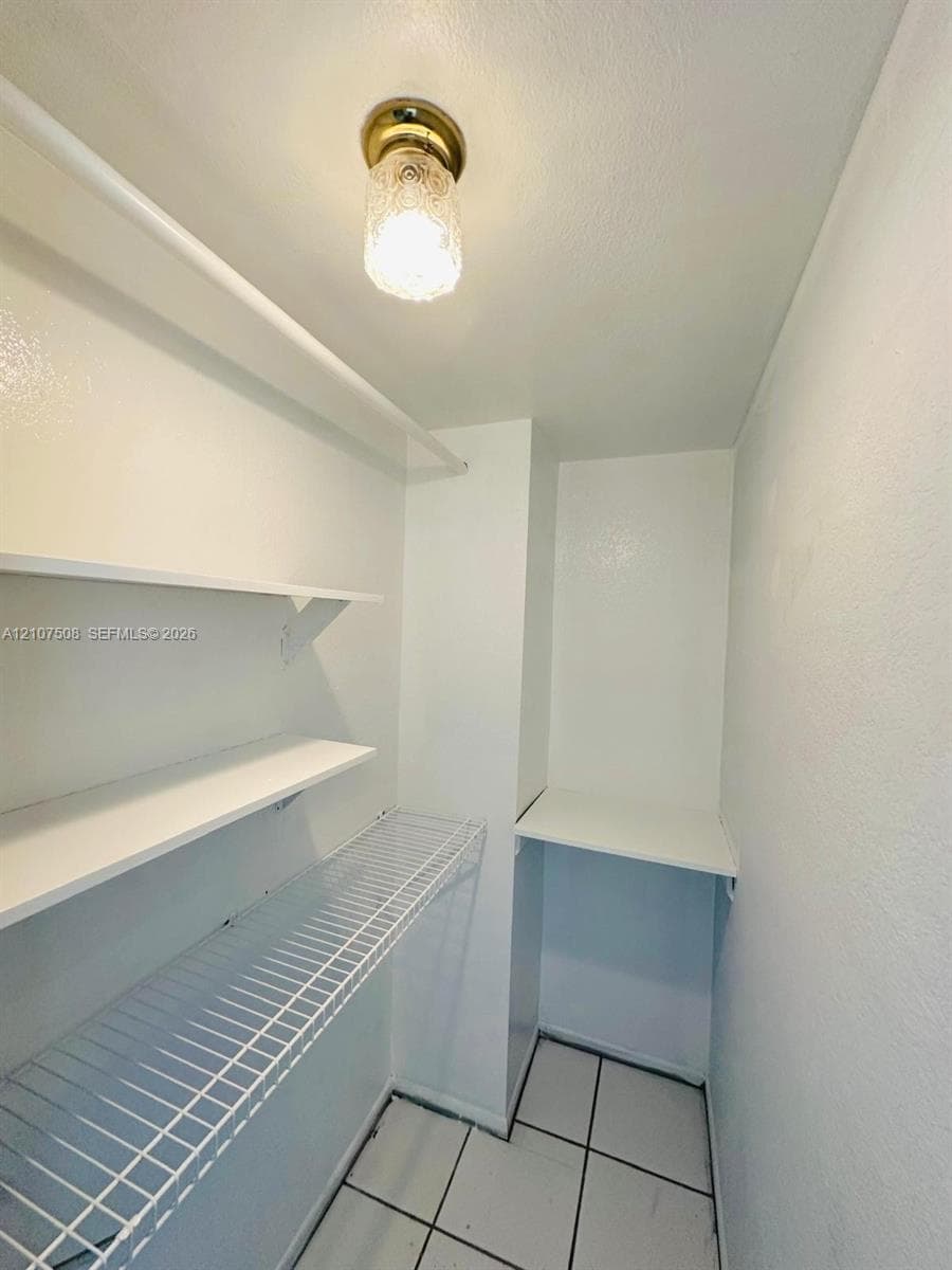 Storage space featuring white solid shelving, white wire shelving, tile flooring, and a flush-mount brass-finish light fixture with textured glass