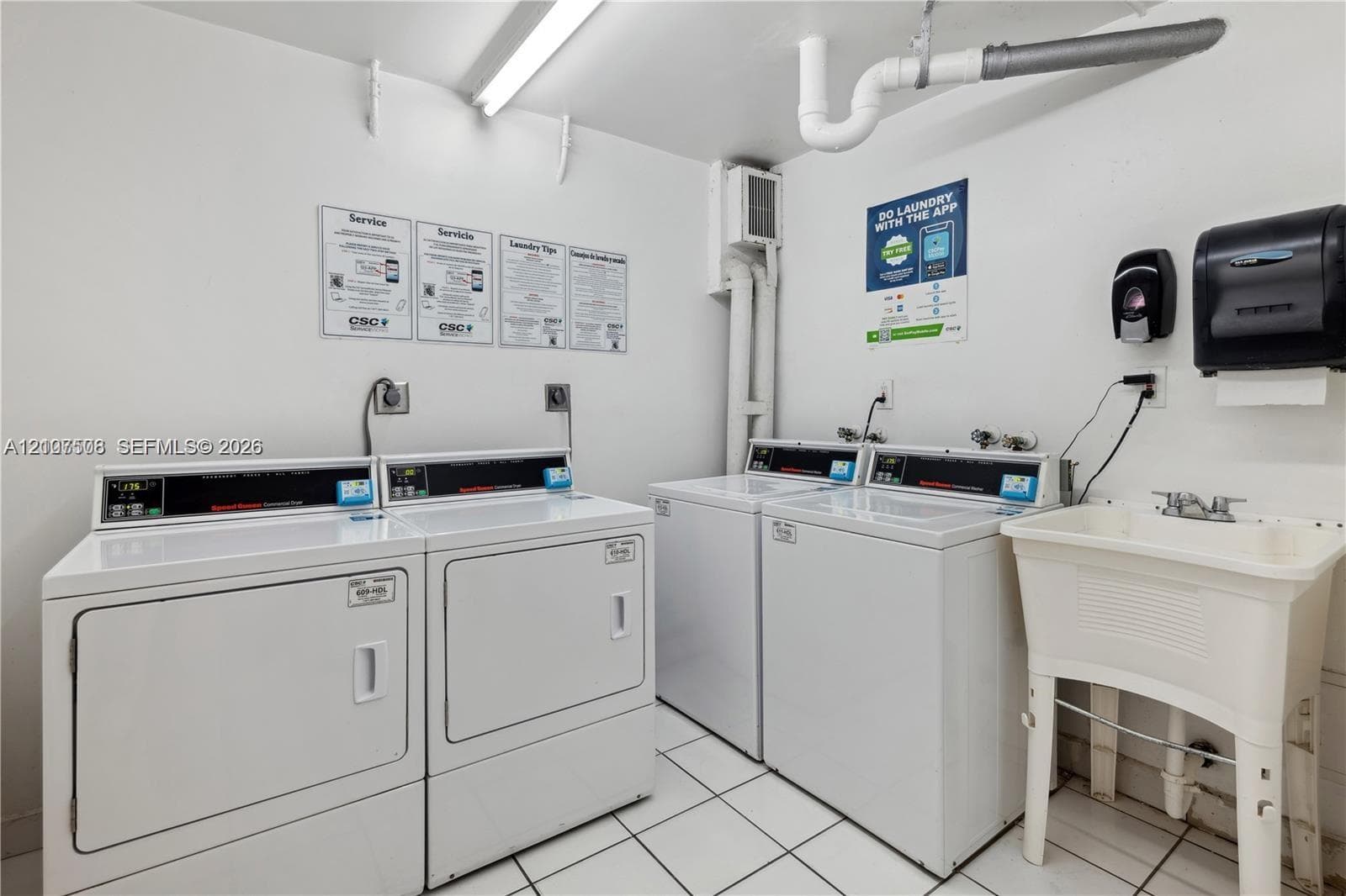 Laundry facility featuring dual white coin-operated washers and dryers, a utility sink with a dual-handle faucet, and white ceramic tile flooring