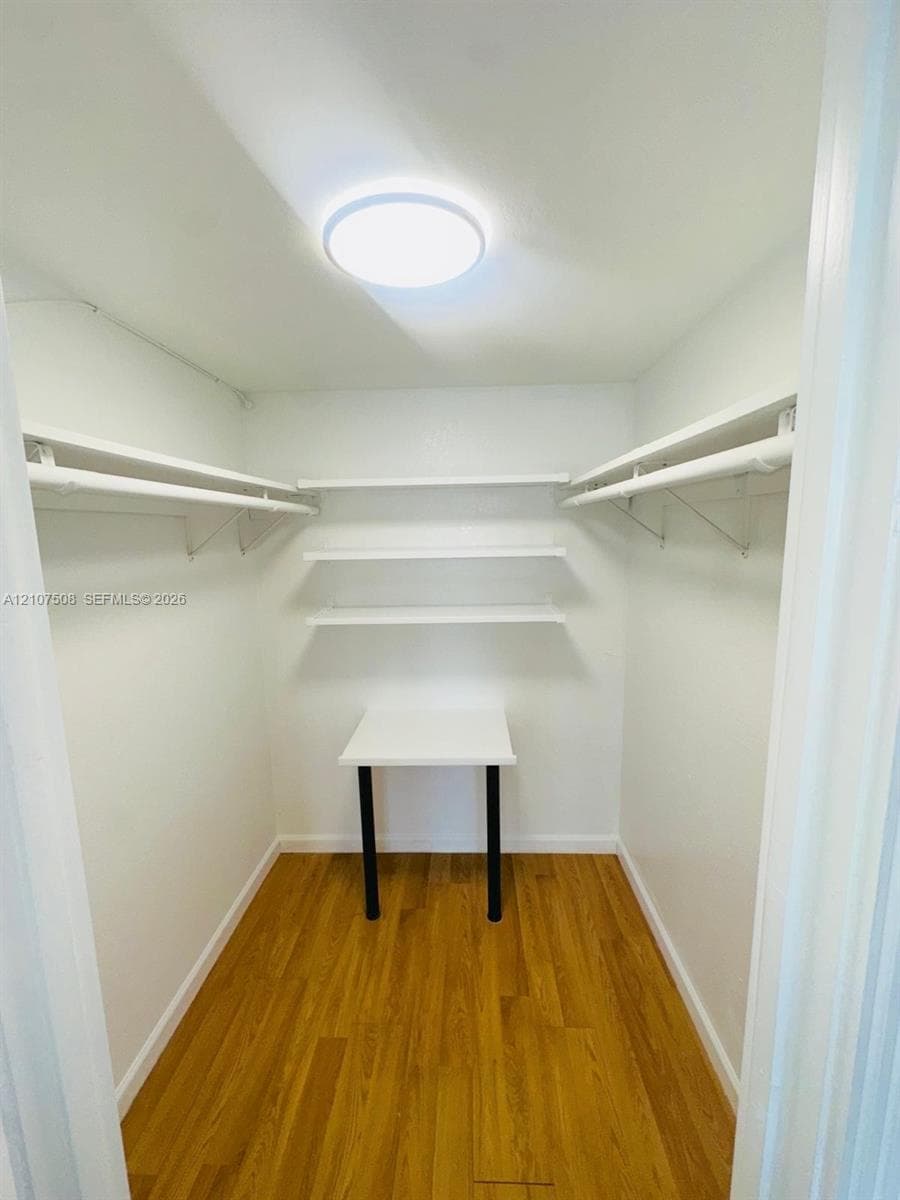 Walk-in closet featuring wood-finish flooring, built-in shelving, integrated clothing rods, a fixed wall-mounted desk, and flush-mount ceiling lighting