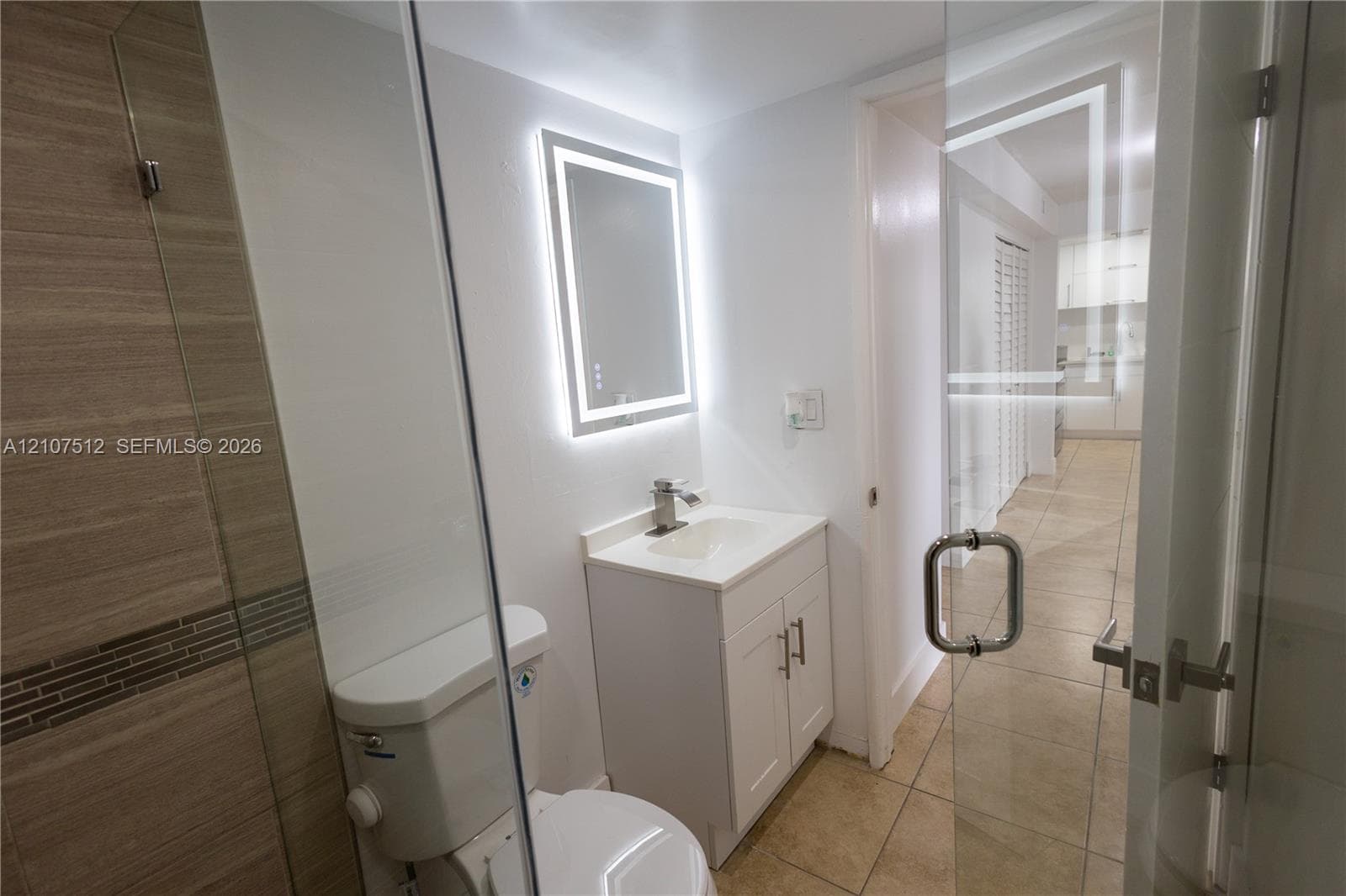 Bathroom featuring a glass-enclosed shower with tiled walls, an illuminated vanity mirror, a white cabinet vanity with a sink, and ceramic tile flooring