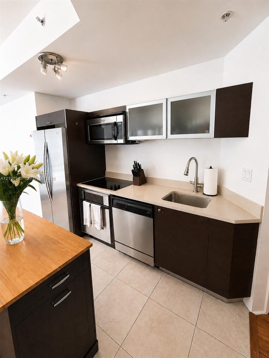 Kitchen featuring dark-finish cabinetry, stainless steel appliances, neutral stone-finish countertops, ceramic tile flooring, and an island with a wood-finish top