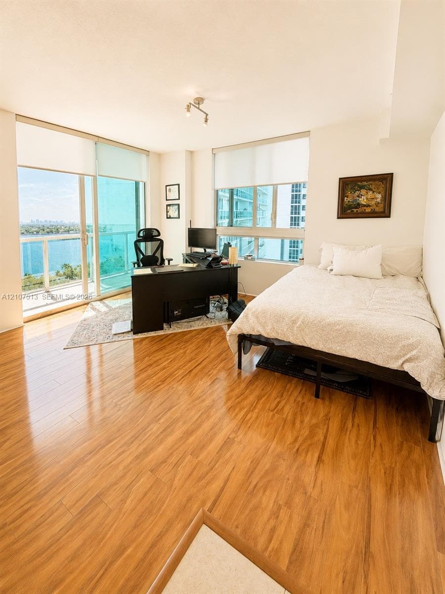 Wood-finish flooring, floor-to-ceiling glass sliding doors, window roller shades, and a glass-paneled balcony railing with water scenery