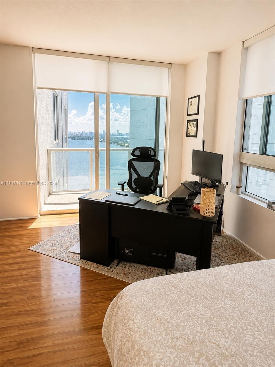 Floor-to-ceiling windows, wood-finish flooring, and a private balcony featuring metal and glass railings with coastal and urban scenery