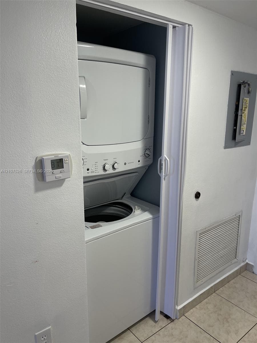 Stacked laundry unit set within a closet, featuring bi-fold doors, wall-mounted digital thermostat, and neutral tile flooring