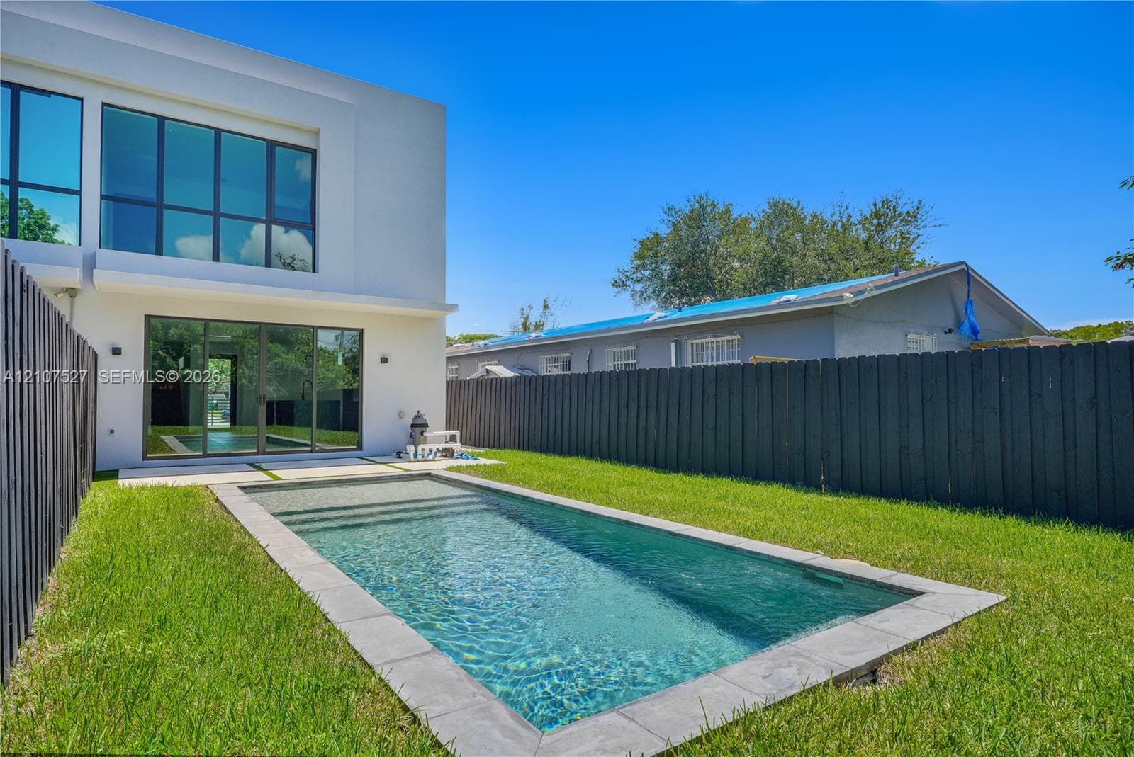 Modern exterior featuring multi-pane glass walls, rectilinear in-ground swimming pool with stone coping, manicured lawn, and perimeter wood privacy fencing
