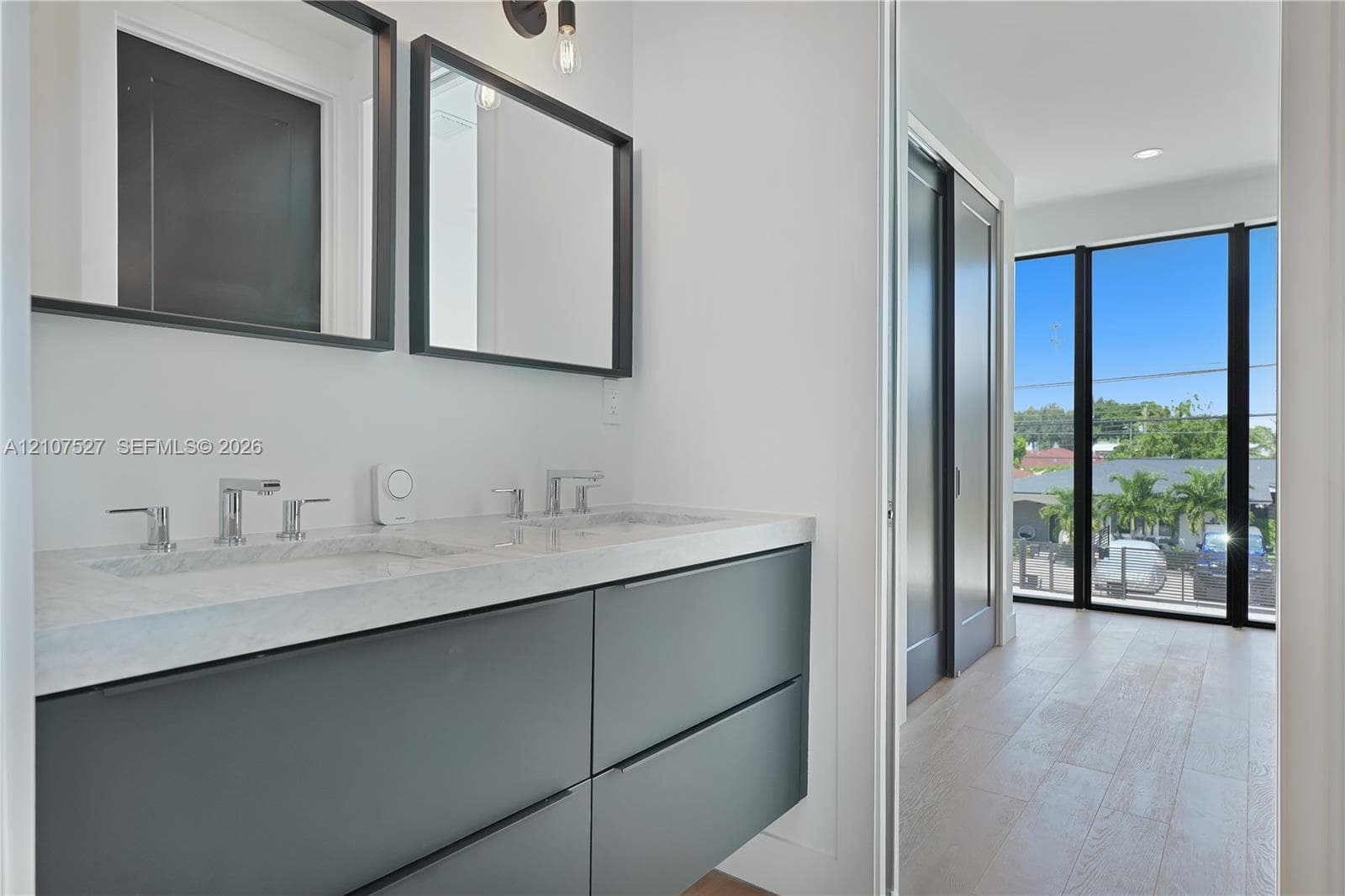 Floating vanity with dual-basin stone countertop, wall-mounted matte black-framed mirrors, wood-finish flooring, and floor-to-ceiling windows