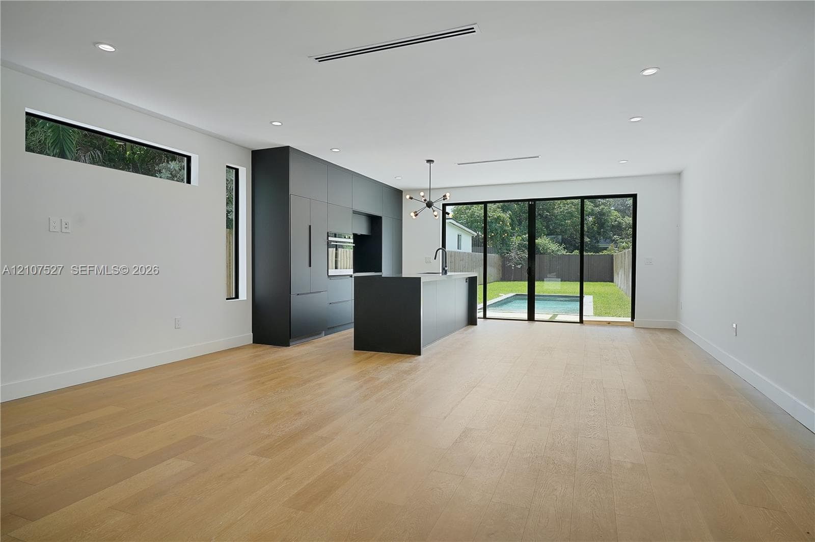 Modern kitchen featuring matte charcoal cabinetry, a central island, wood-finish flooring, recessed lighting, and multi-panel glass sliding doors opening to the exterior pool area