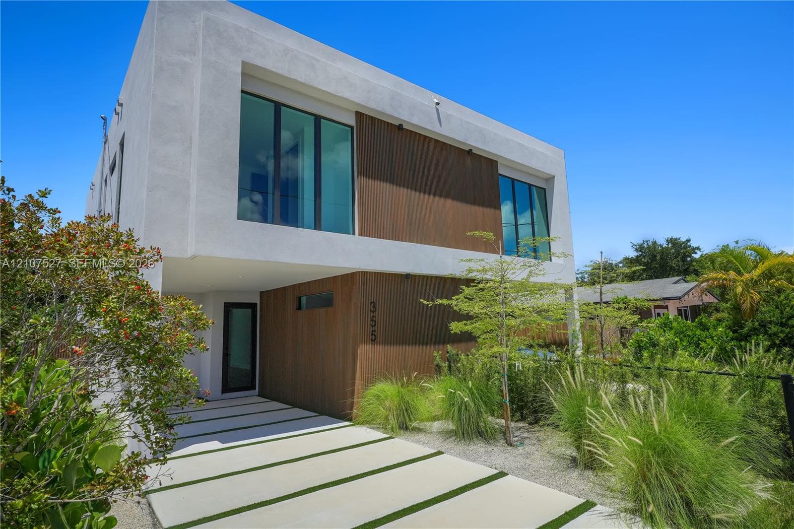 Modern two-story architectural exterior featuring smooth white stucco finish, vertical wood-finish siding, oversized floor-to-ceiling windows, and a segmented concrete paver driveway with integrated turf strips
