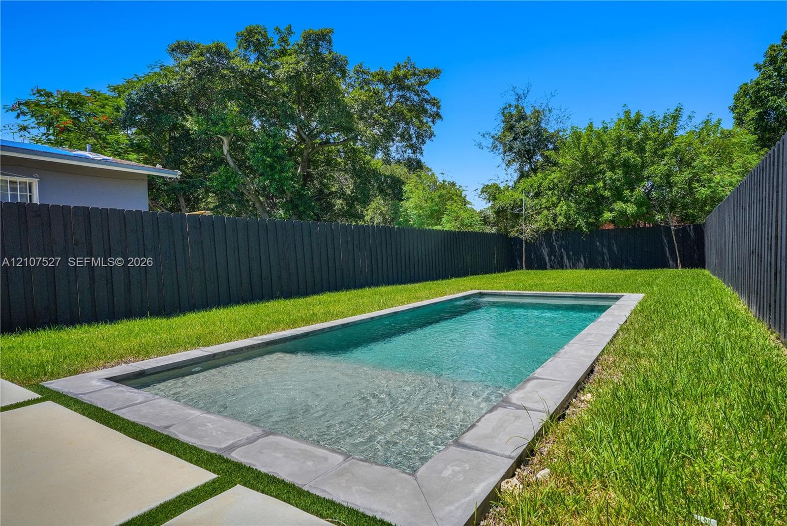 Rectangular swimming pool featuring stone coping, integrated shallow shelf, manicured lawn, and full-perimeter privacy fencing