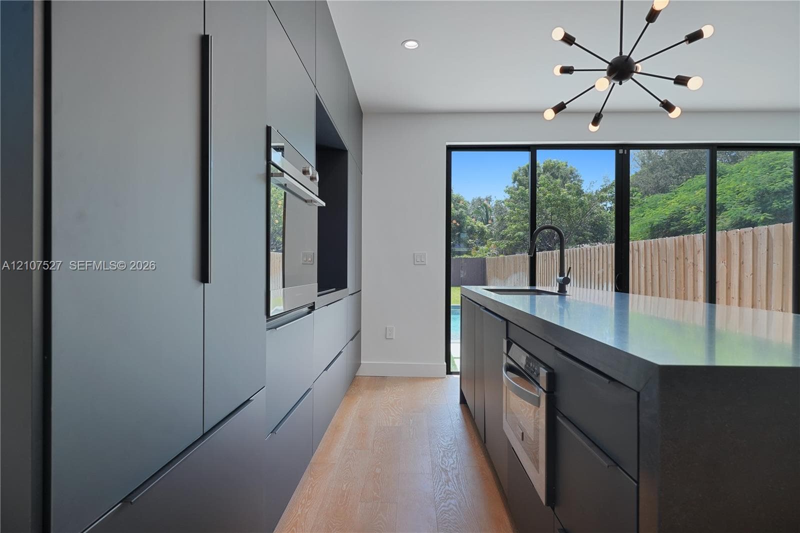 Modern kitchen featuring charcoal slab-front cabinetry, an expansive island with a matte black faucet, integrated stainless steel appliances, wood-finish flooring, and multi-panel glass doors