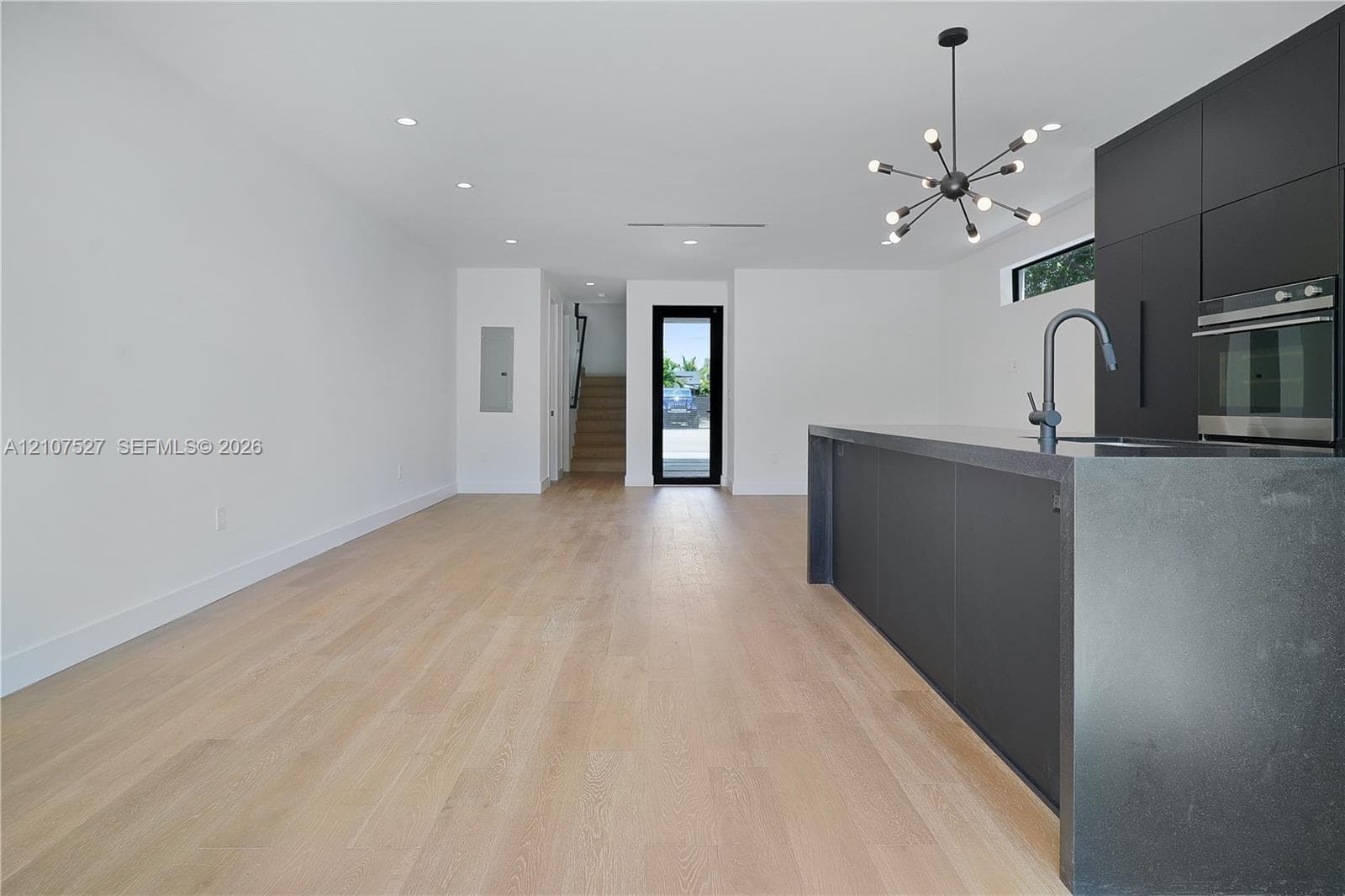 Modern kitchen featuring matte black flat-panel cabinetry, a dark waterfall-edge island, recessed lighting, a multi-light contemporary chandelier, and light-toned wood-finish flooring