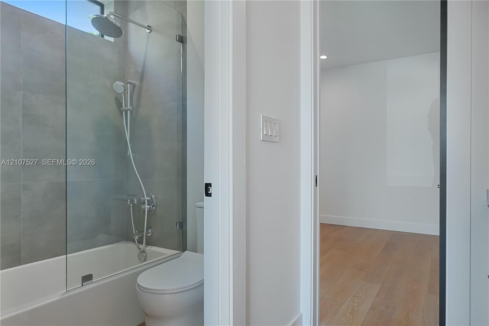 Bathroom featuring large-format grey tile surround, glass panel shower screen, rainfall shower head, and wood-finish flooring in the room beyond