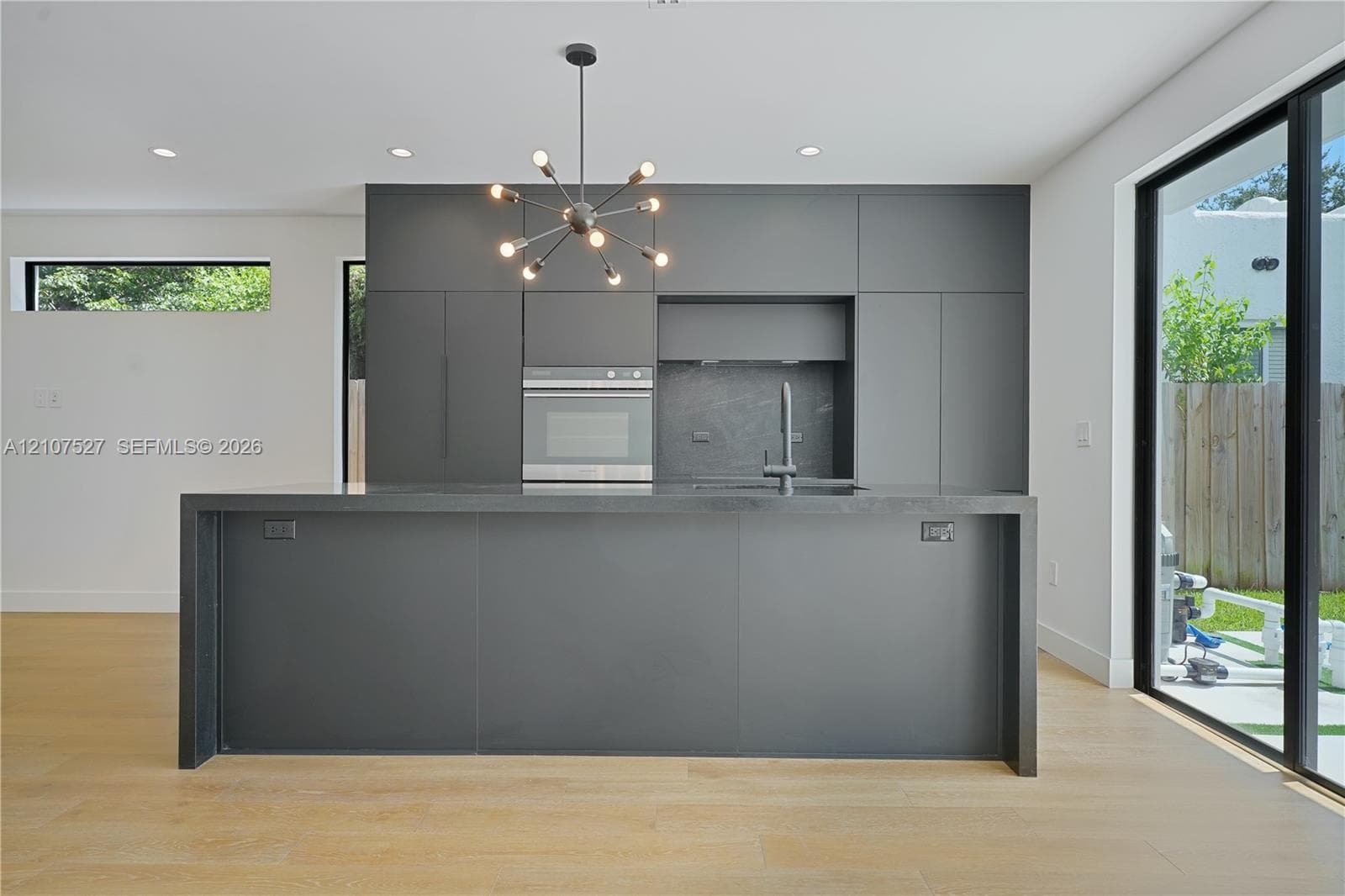 Kitchen featuring matte charcoal flat-panel cabinetry, a central island with a waterfall countertop, integrated stainless steel oven, multi-arm chandelier, and wood-finish flooring