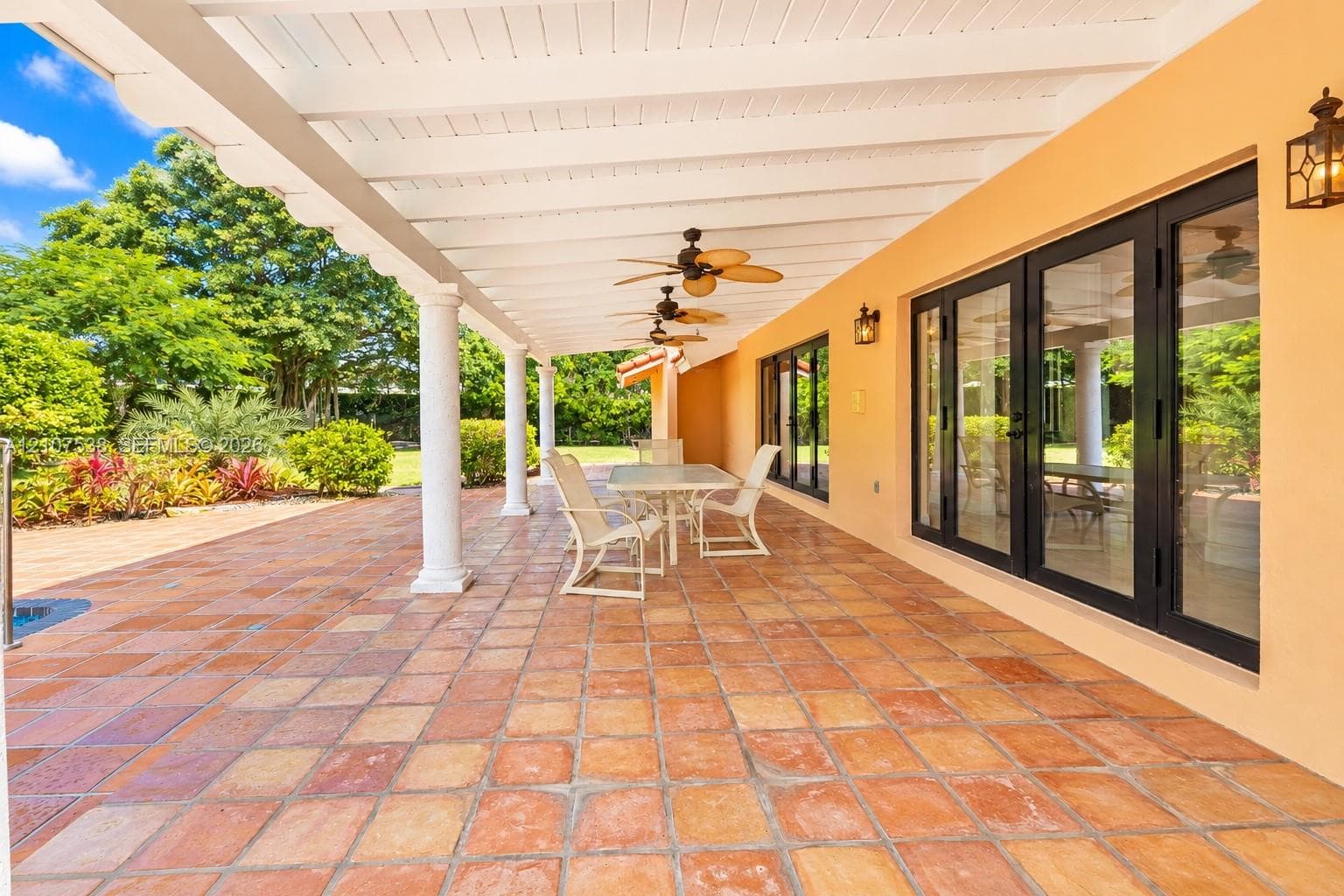 Covered lanai featuring terracotta-tile flooring, white structural columns, ceiling-mounted fans, and black-framed multi-panel glass doors