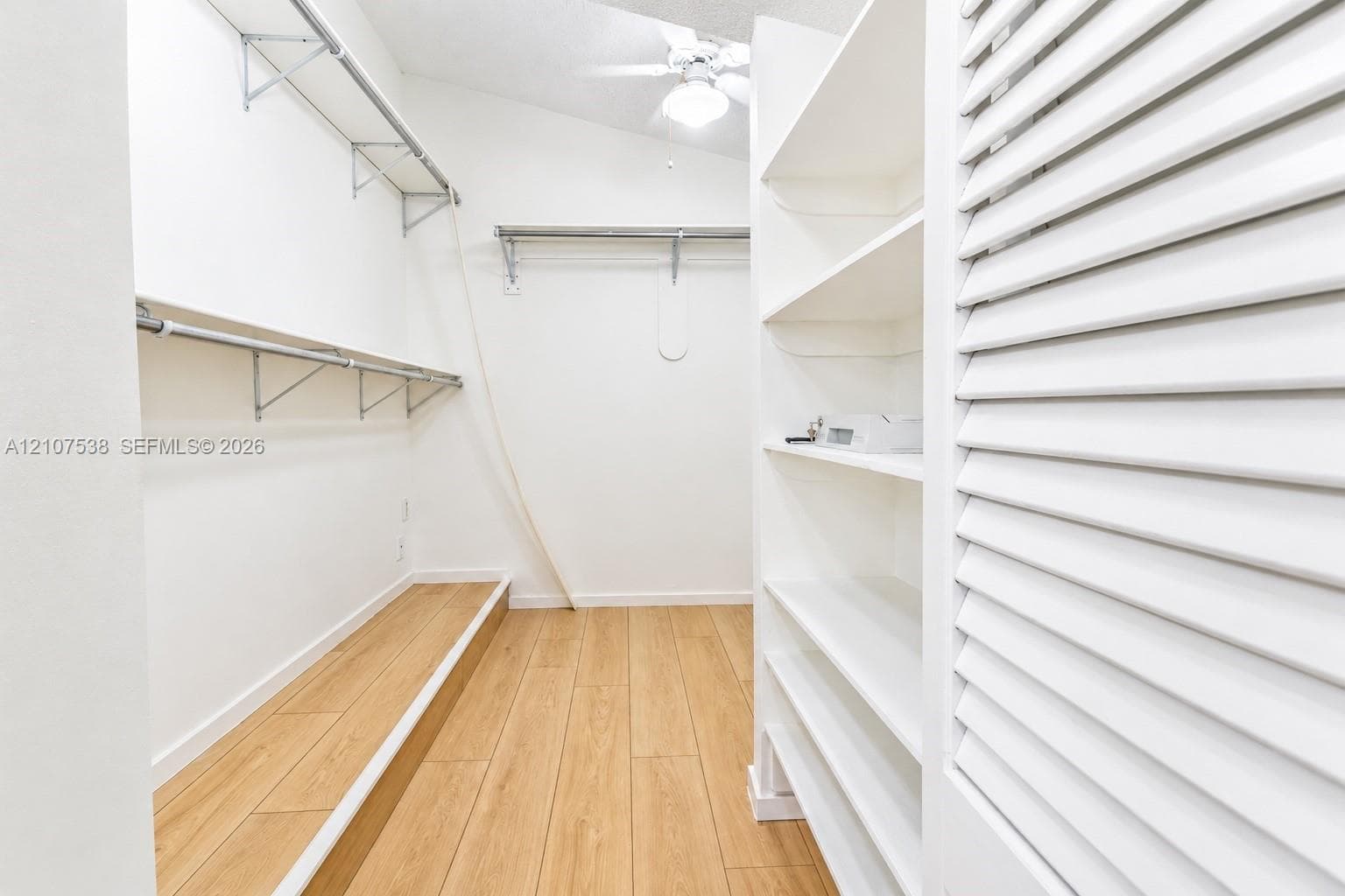 Walk-in closet featuring wood-finish flooring, built-in shelving, metal garment rods, louvered closet doors, and an overhead ceiling fan with lighting