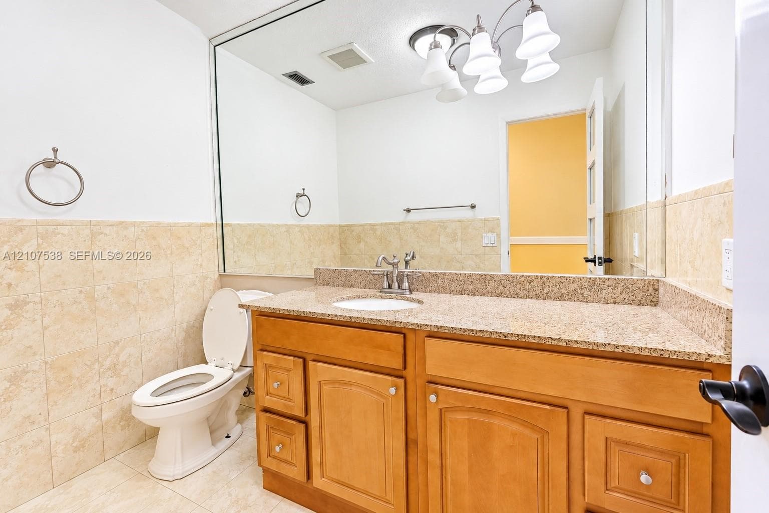 Bathroom featuring a vanity with wood cabinetry, a stone-finish countertop with an integrated basin, a full-width mirror, tile wainscoting, and a multi-light fixture