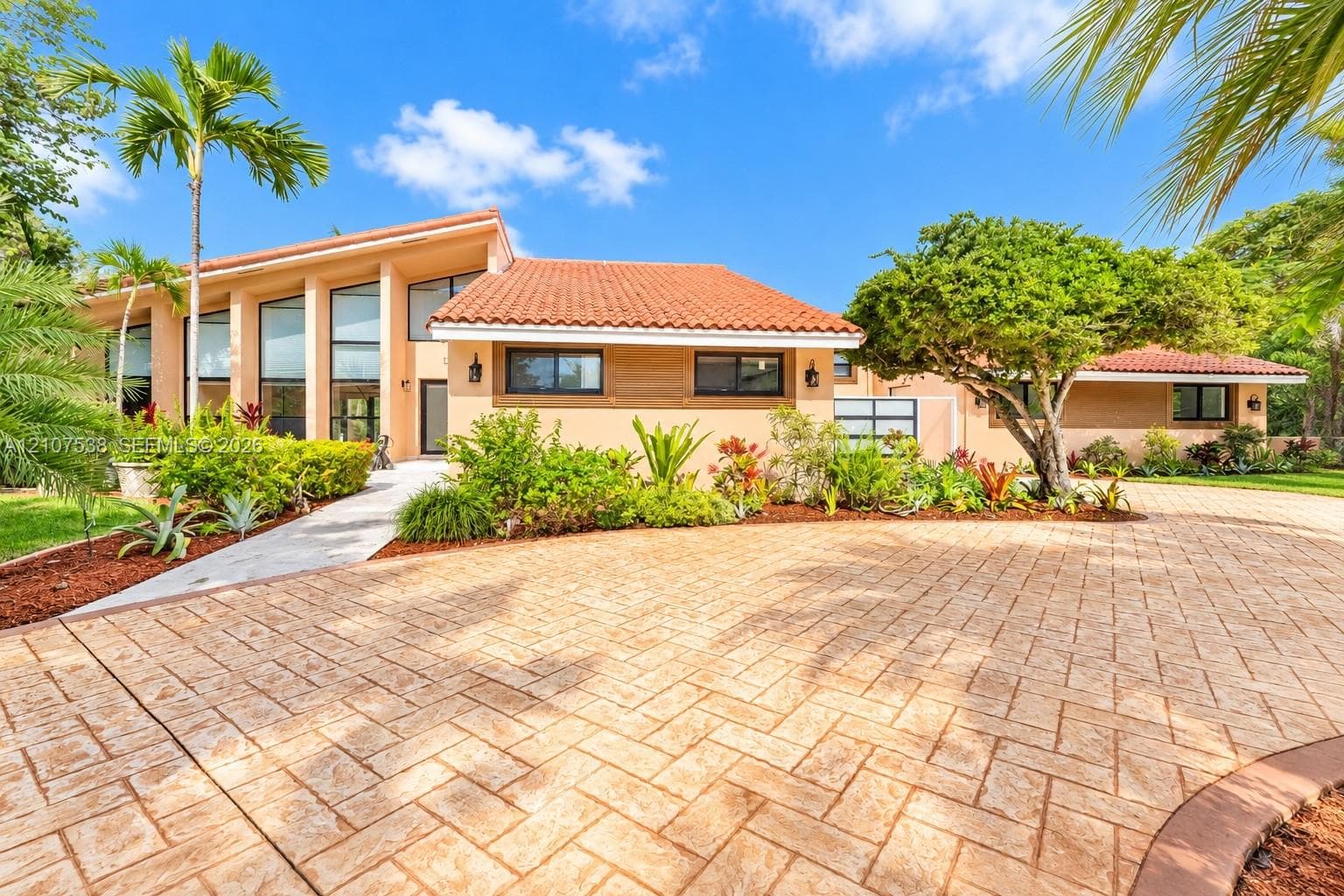 Stucco exterior featuring a terracotta tile roof, expansive floor-to-ceiling windows, patterned paver driveway, and mature tropical landscaping