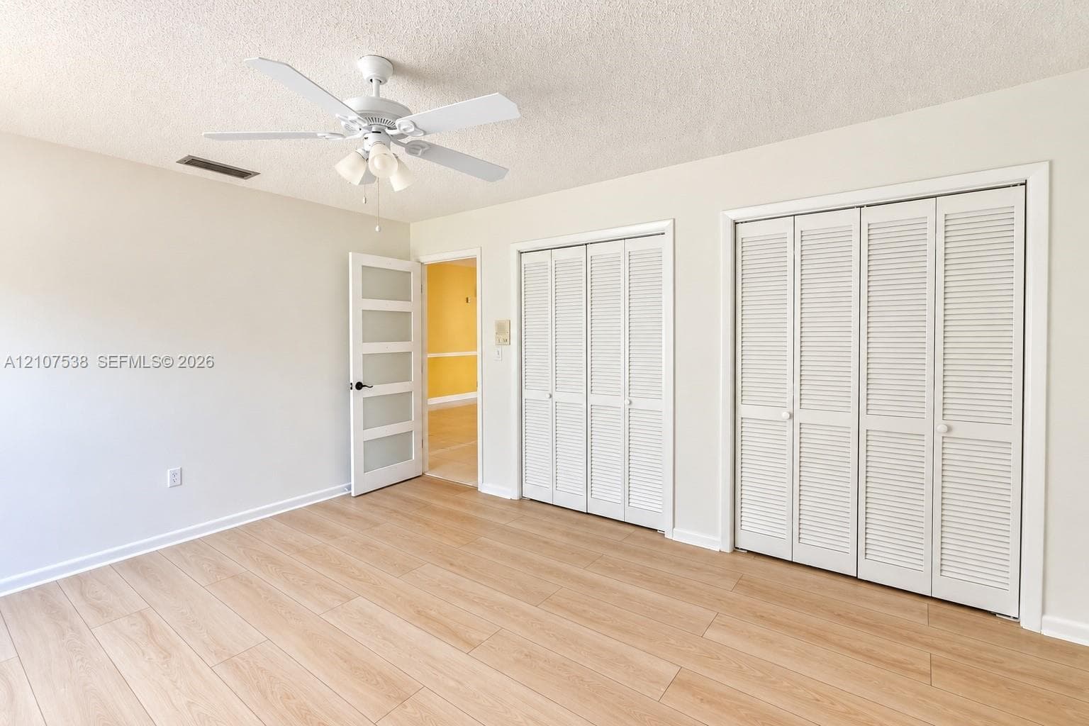 Room featuring wood-finish flooring, dual closets with louvered doors, a five-panel frosted glass door, and an overhead ceiling fan with integrated lighting
