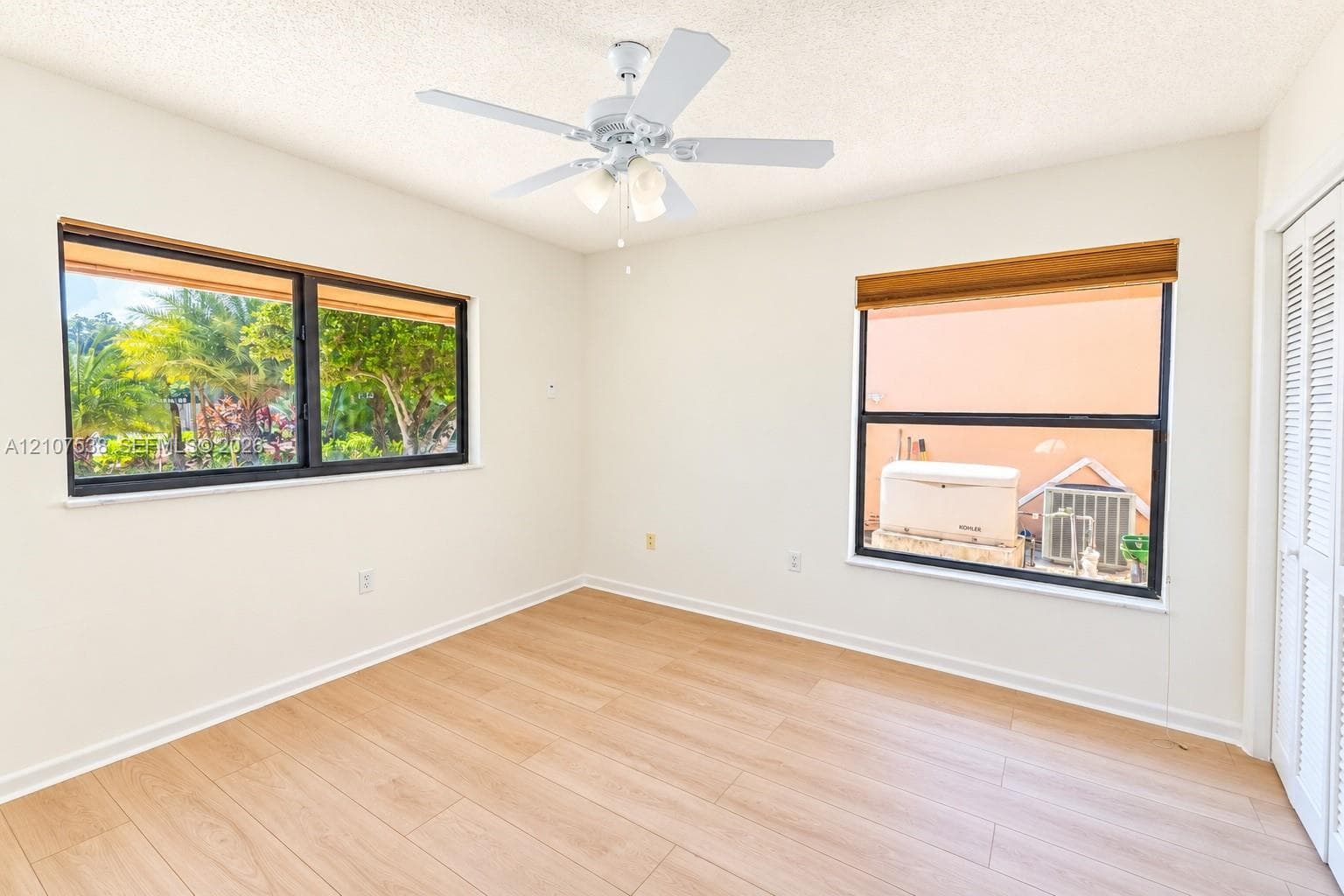 Interior space featuring light-toned wood-finish flooring, dual windows with black frames, white louvered closet doors, and a ceiling fan with integrated lighting
