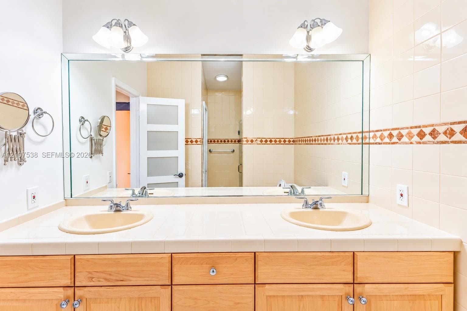 Dual-sink vanity with a tile countertop, large wall-mounted mirror, light wood cabinetry, and decorative tile backsplash