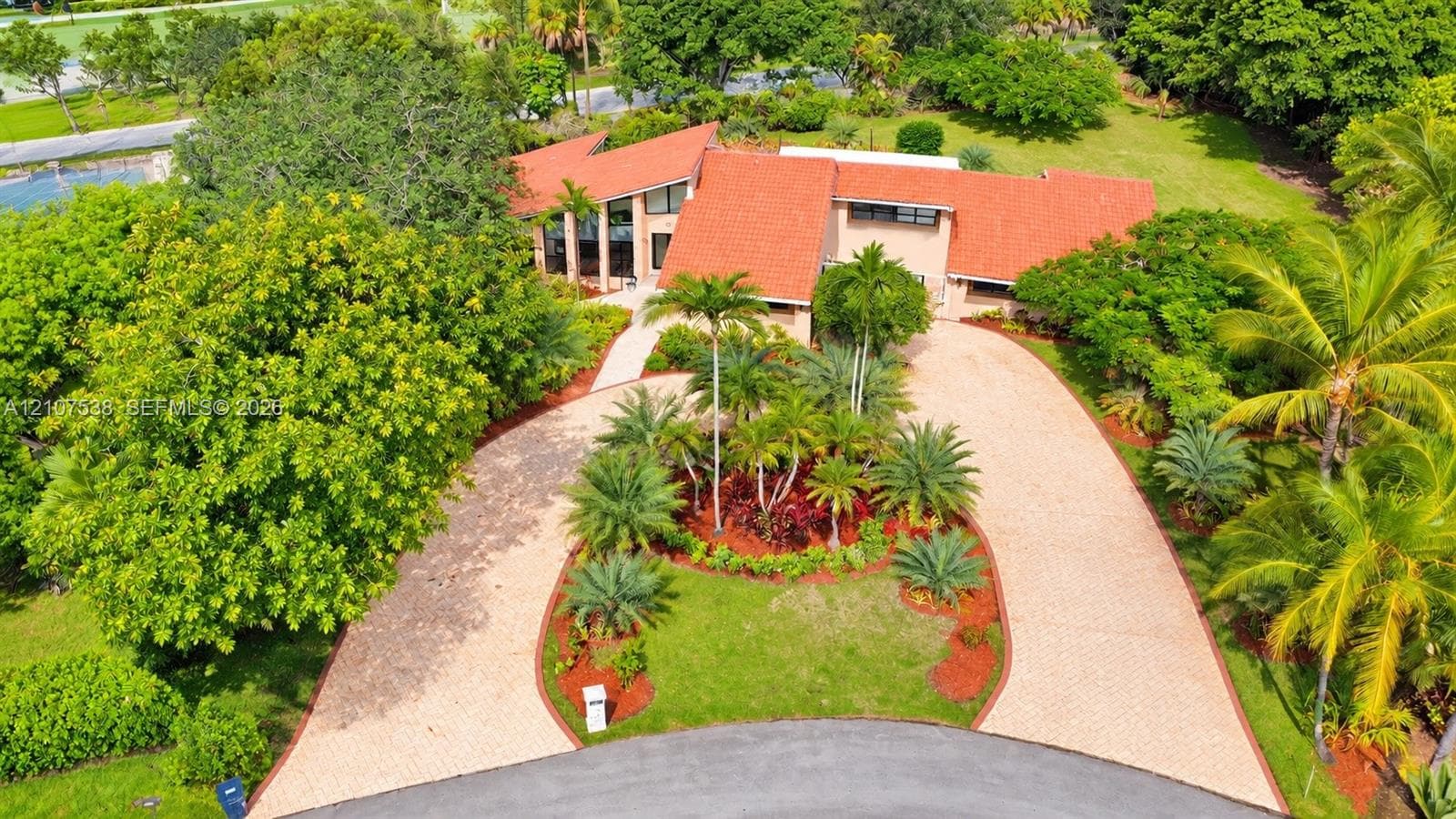 Multi-level residence featuring a textured clay tile roof, expansive circular paver driveway, manicured landscaping, and mature foliage