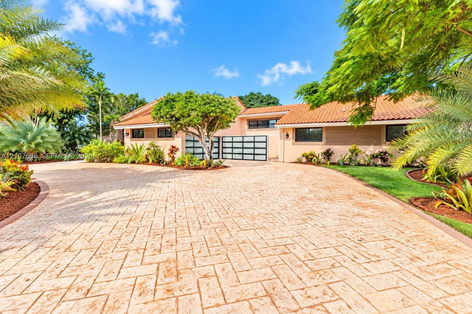 Stamped concrete driveway featuring a herringbone pattern, terracotta roof tiles, and modern garage doors with frosted glass panels, accented by mature palm trees and manicured landscaping