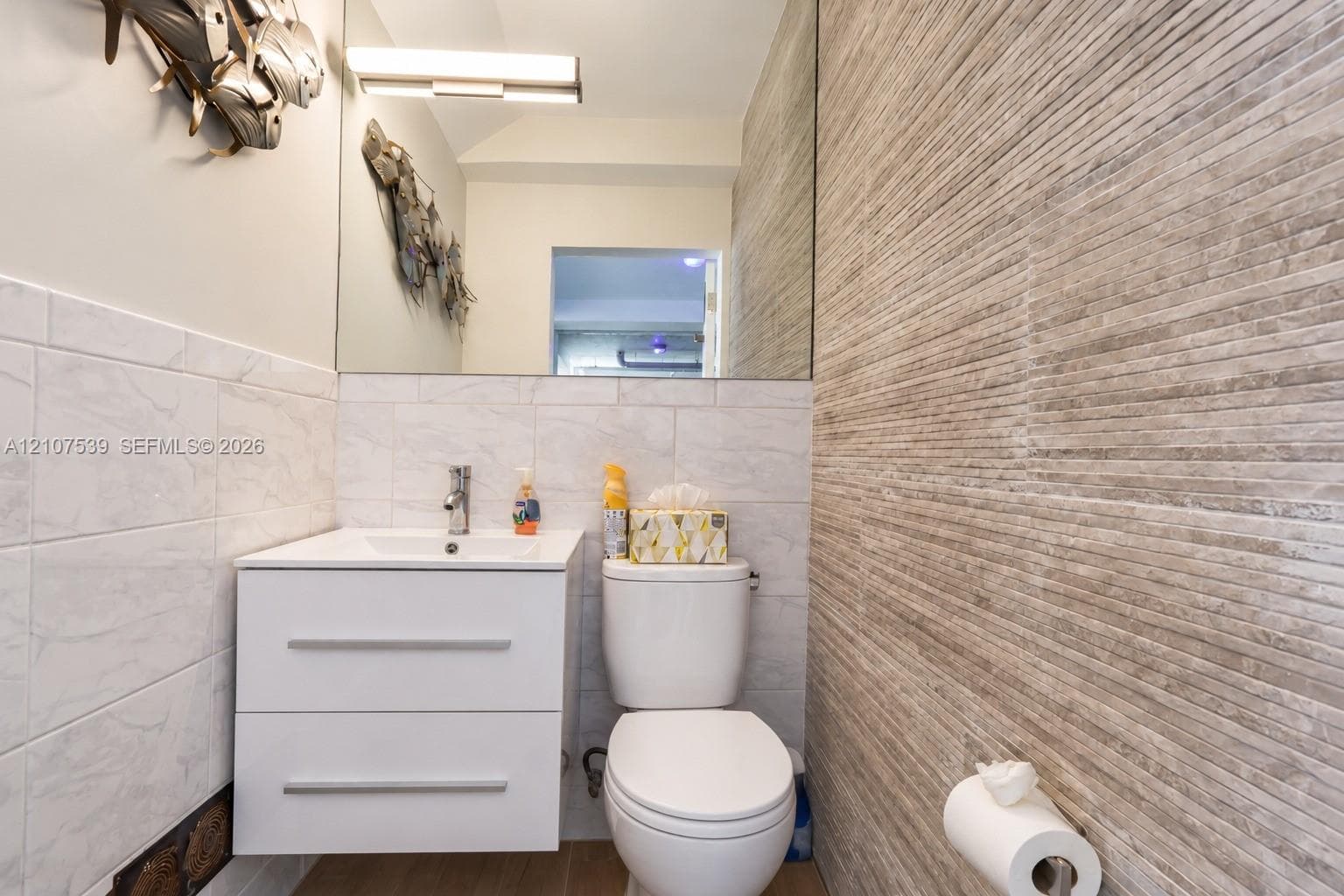 Floating white vanity with integrated sink, textured stone-finish accent wall, large format light-colored wall tiling, wall-mounted vanity lighting, and frameless mirror