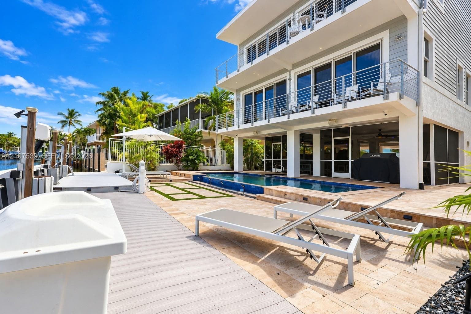 Three-story waterfront residence featuring a multi-level balcony system with cable railings, a private in-ground swimming pool, stone-paver hardscaping, and a synthetic deck area with a boat dock