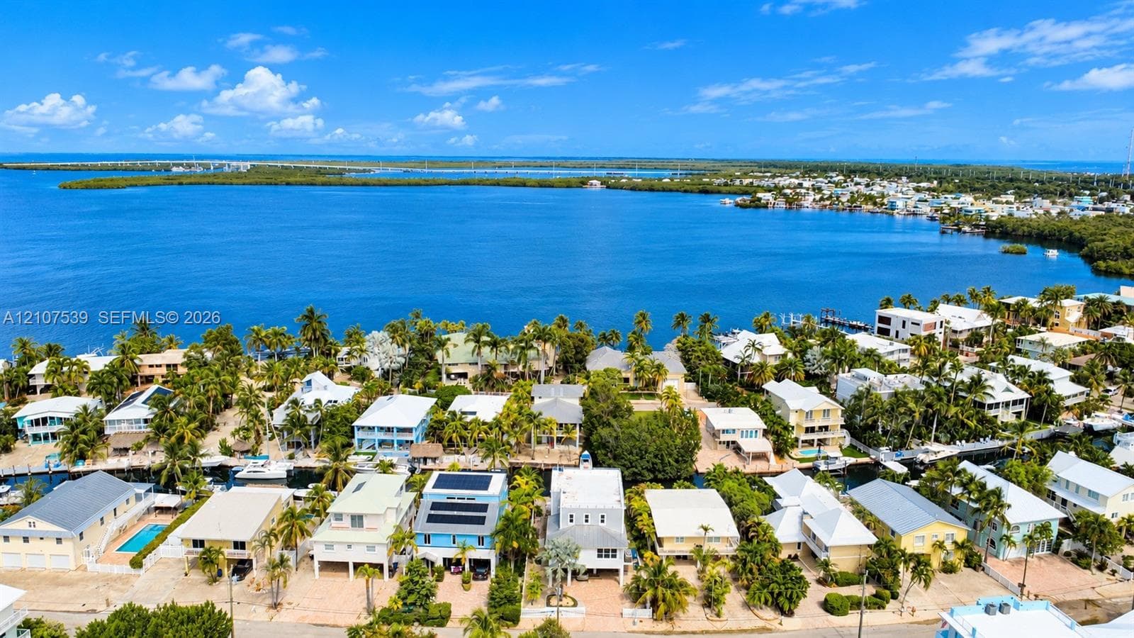 Aerial perspective of coastal residential property featuring canal-front access, paved driveways, elevated structural design, and solar panel arrays, set against a bay backdrop