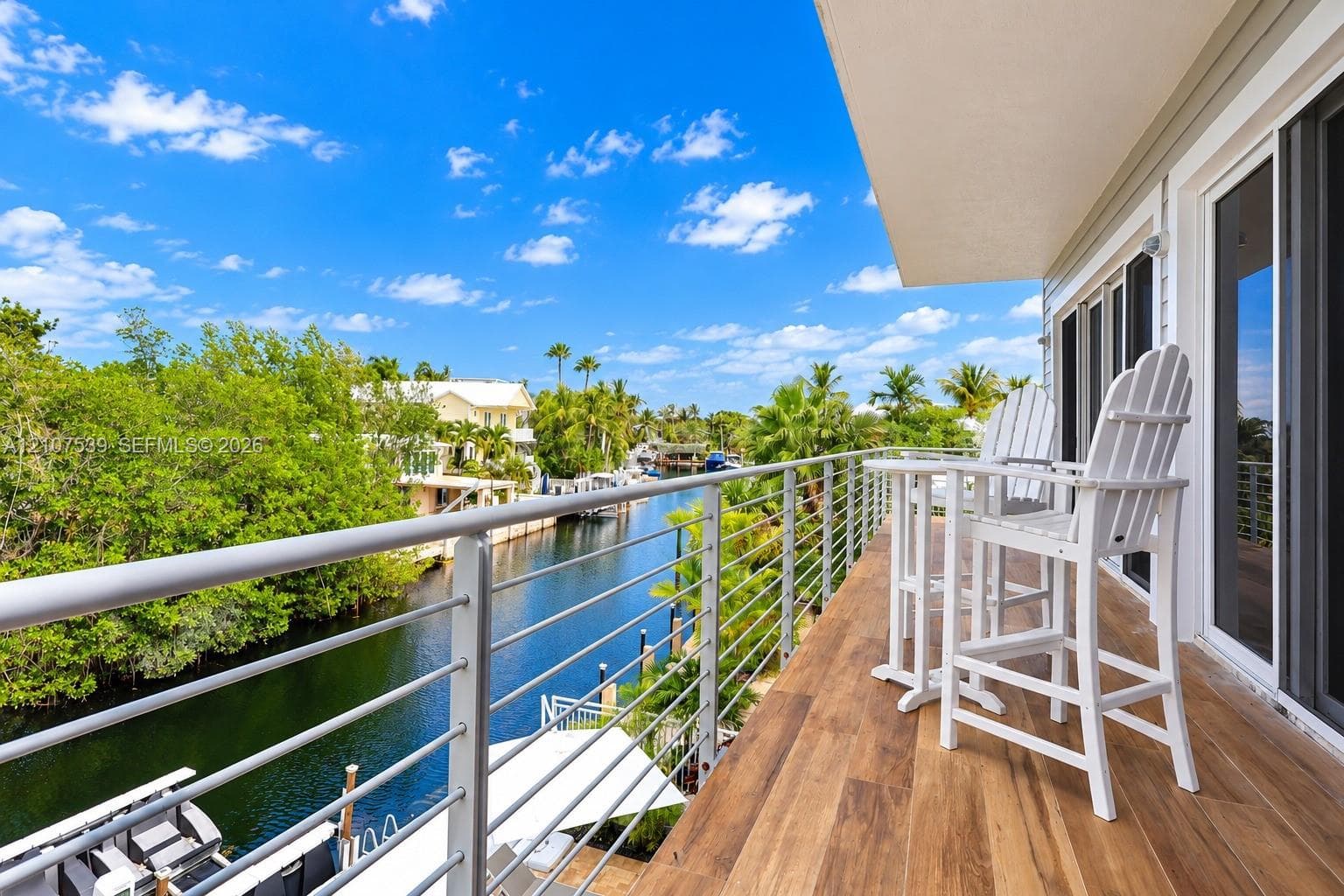 Covered balcony featuring wood-finish flooring, metal cable railing, and sliding glass door access, overlooking a canal and mature landscaping