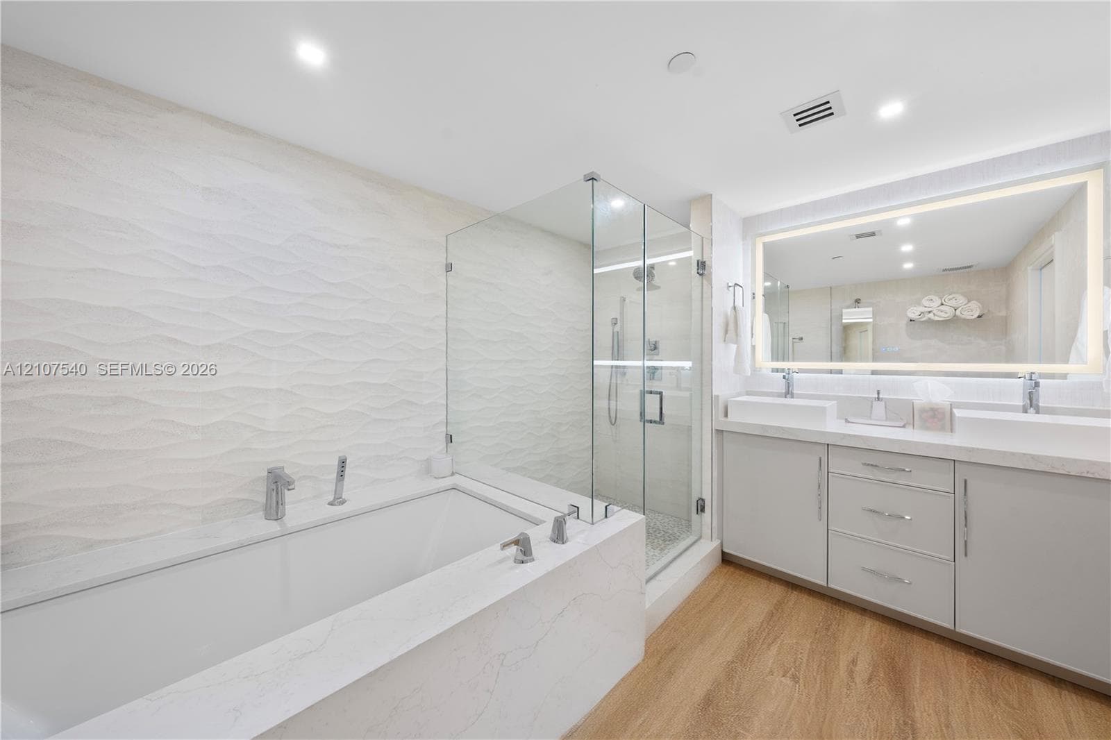 Master bathroom featuring a frameless glass-enclosed shower, a deep soaking tub with stone-finish surround, a dual-sink vanity with comfort height cabinetry, and an oversized back illuminated wall mirror