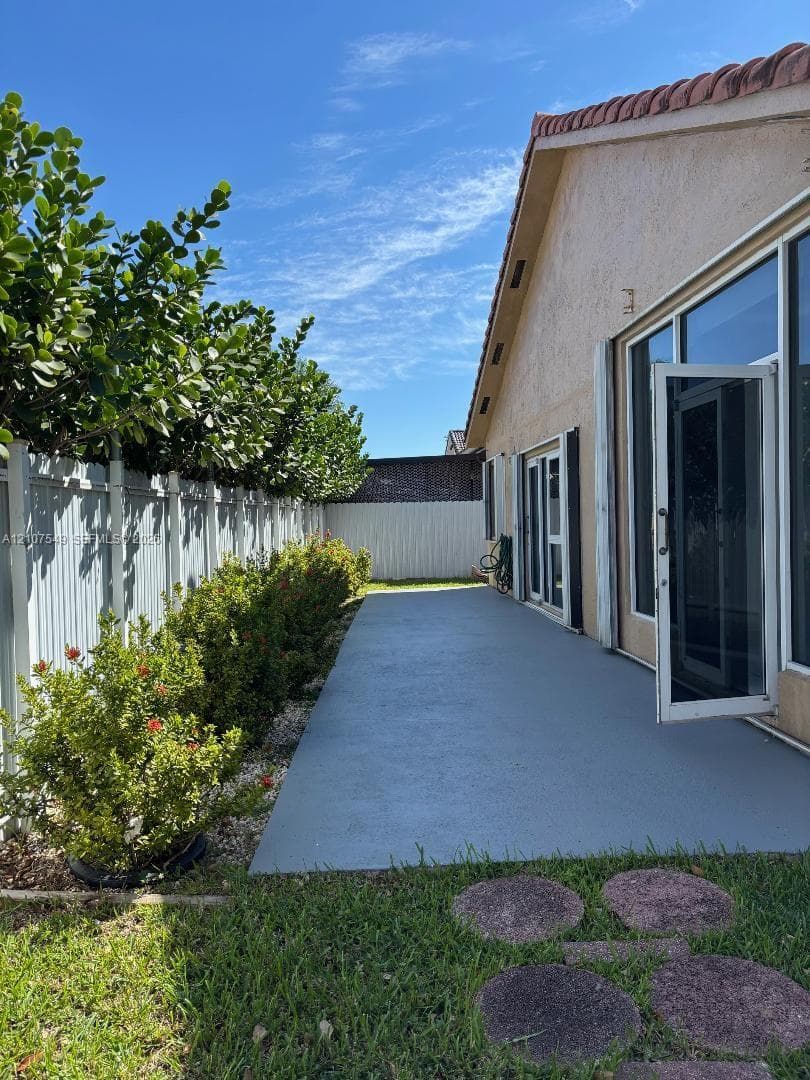 Exterior patio space featuring a concrete surface, perimeter vinyl fencing, mature hedge landscaping, and oversized glass sliding doors