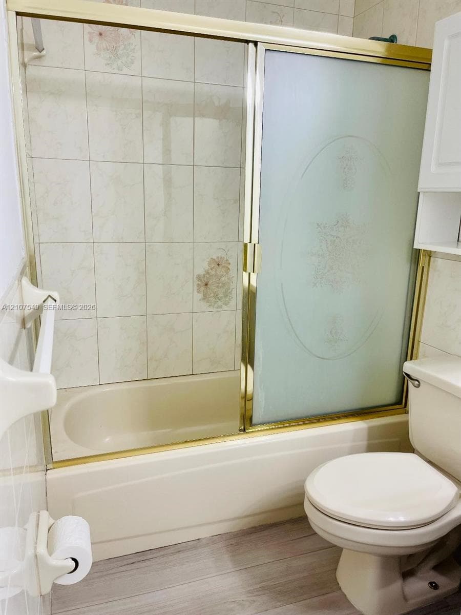 Bathtub with gold-tone frame sliding glass enclosure, decorative tile surround, standard-height toilet, and built-in white wall cabinetry