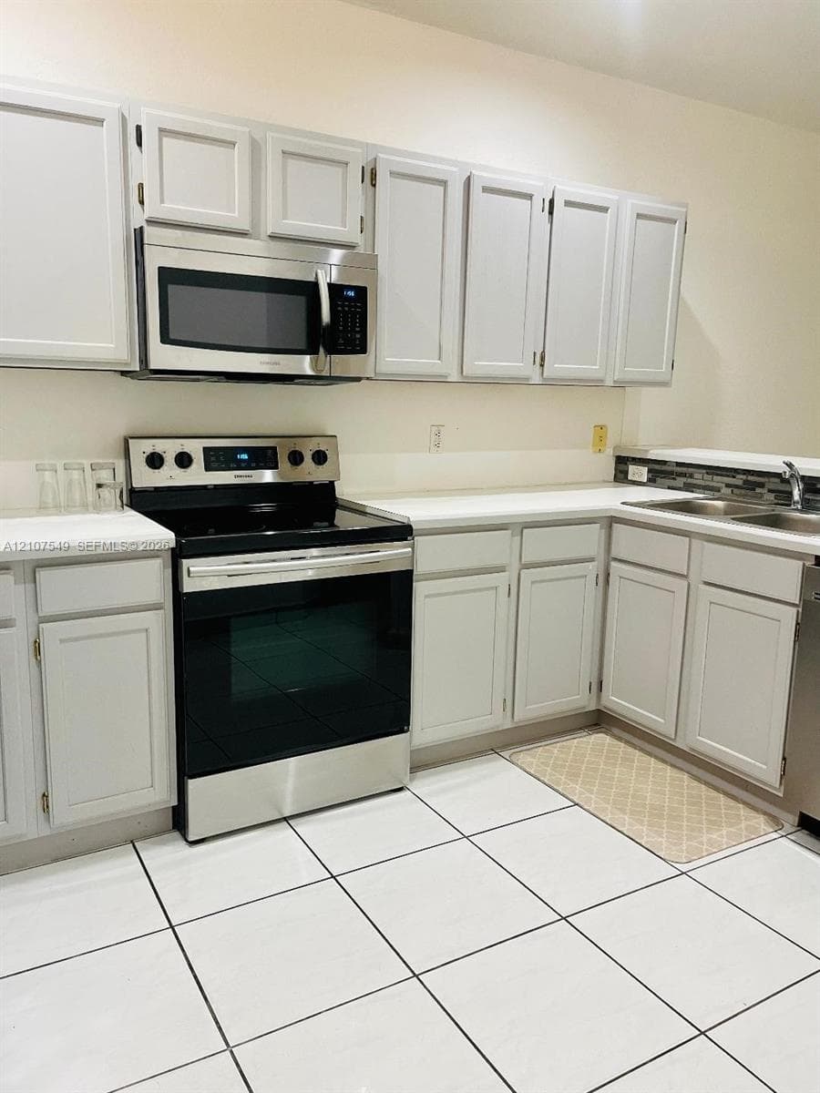 Kitchen featuring white shaker-style cabinetry, stainless steel appliances, solid surface countertops, a mosaic tile backsplash, and ceramic tile flooring