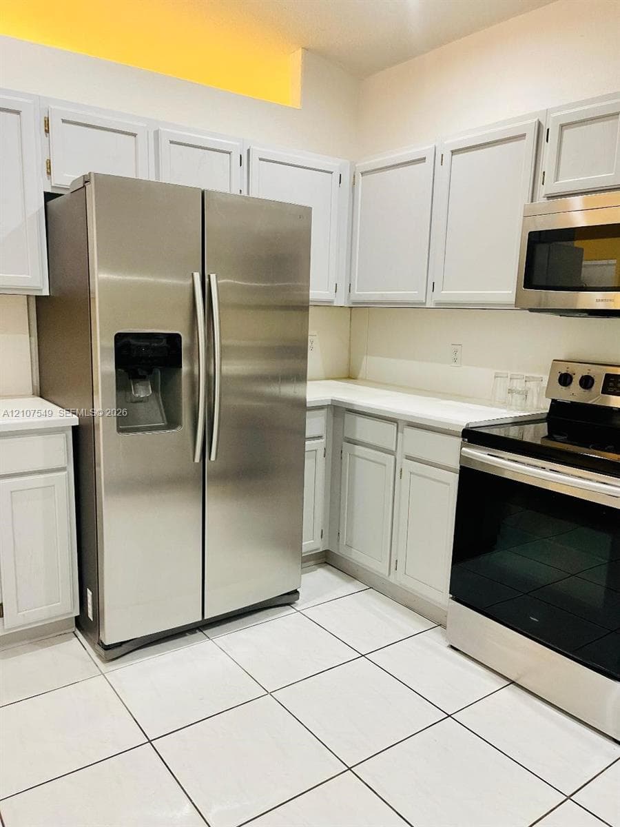Kitchen featuring stainless steel side-by-side refrigerator, microwave, and electric range, white cabinetry, solid surface countertops, and ceramic tile flooring