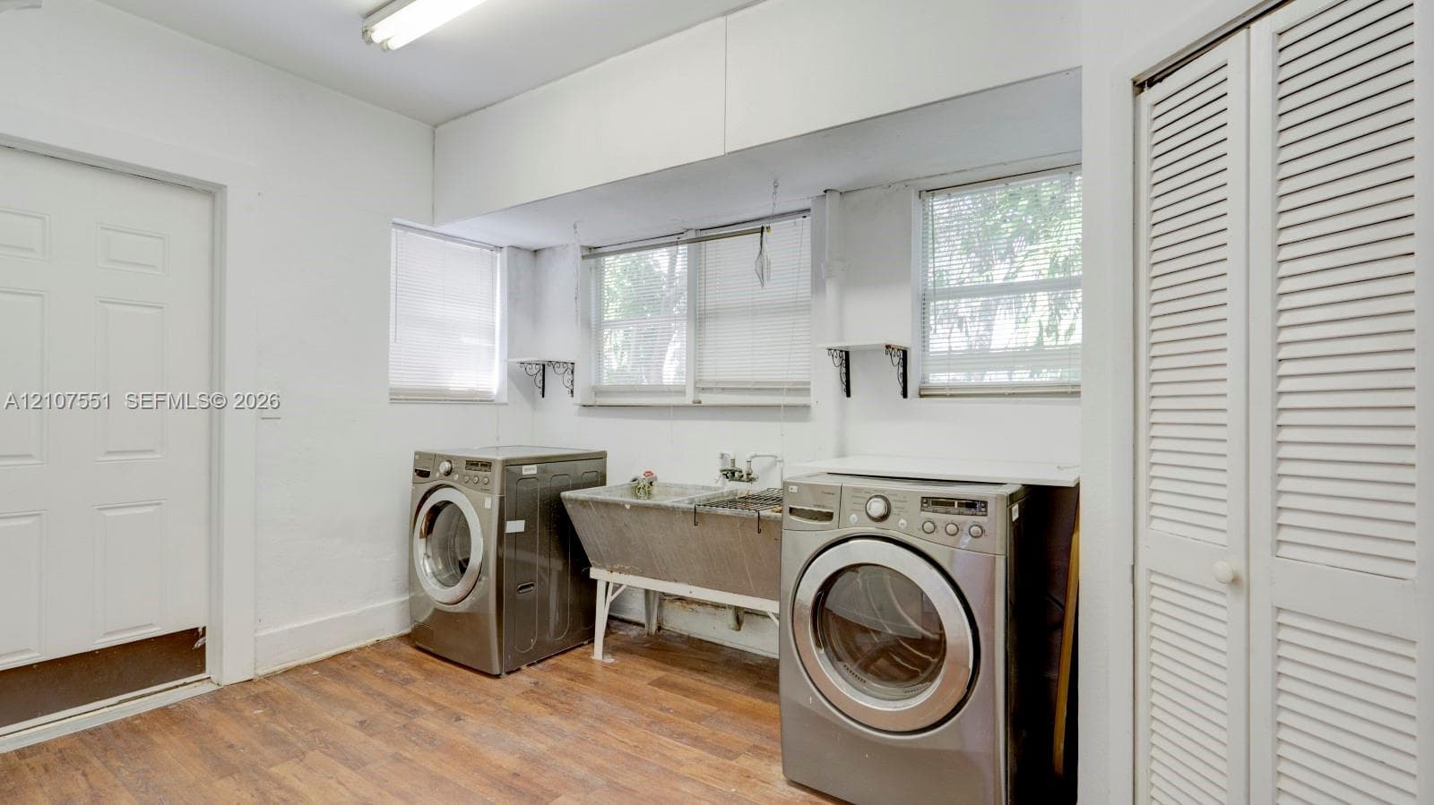 Laundry area featuring wood-finish flooring, a stainless steel utility sink, and louvered closet doors