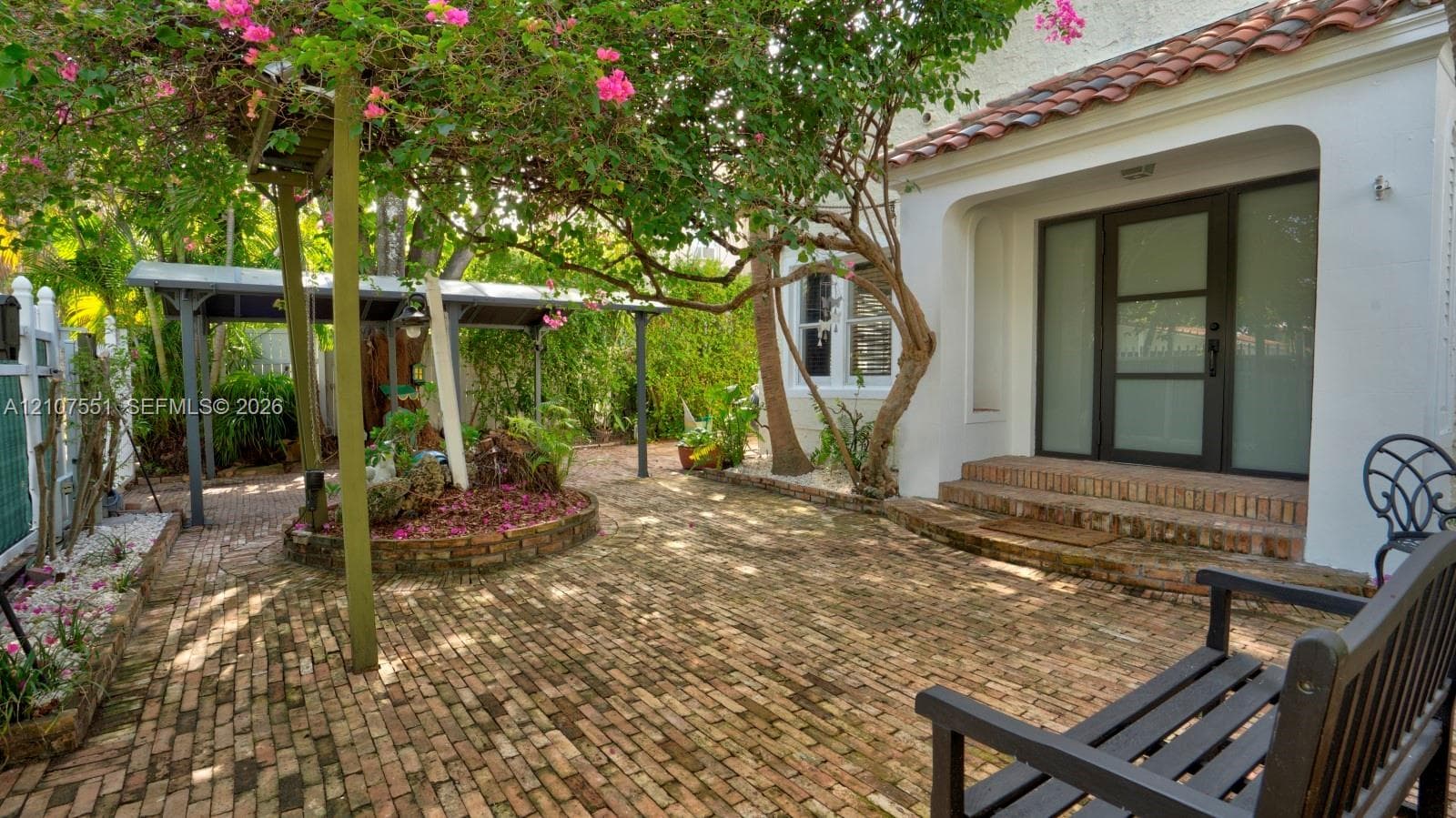 Courtyard entry featuring a herringbone-patterned brick patio, white stucco exterior, terracotta-tiled roof accents, a covered parking structure, and a glass-paneled entry door