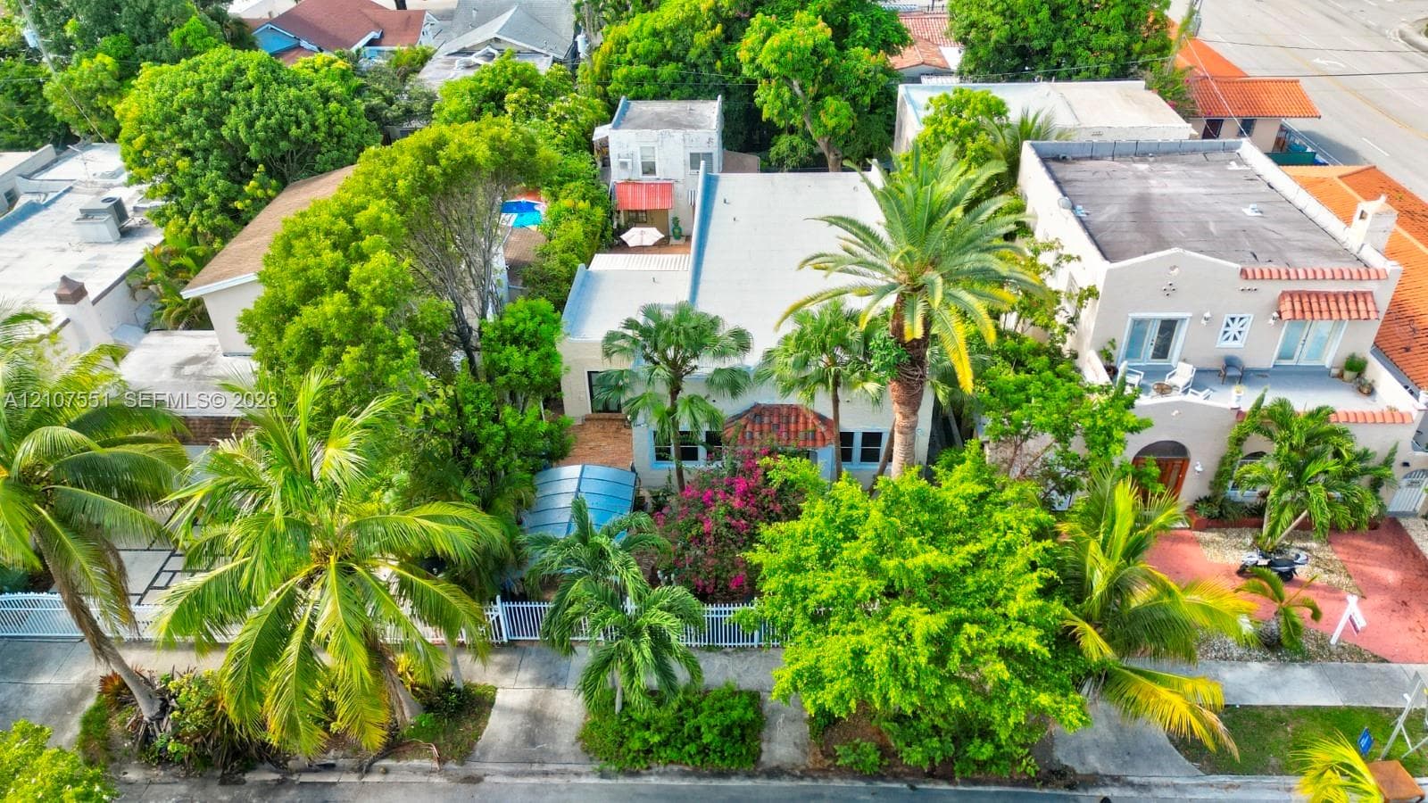 Aerial perspective of a property featuring mature tropical landscaping, palm trees, a concrete-surfaced driveway, and a fenced perimeter