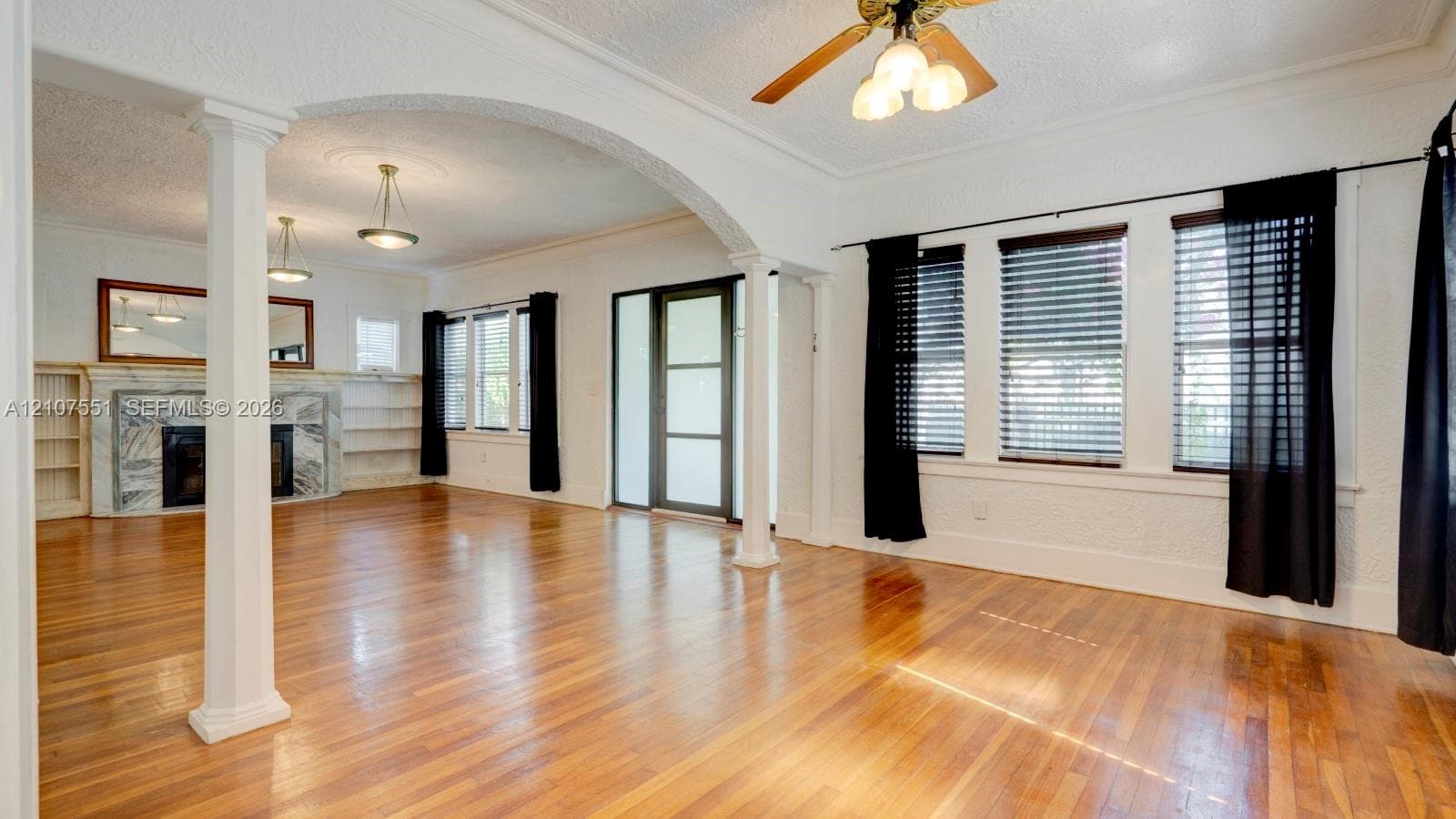 Open living area featuring wood-finish flooring, a stone-surround fireplace, built-in shelving, decorative columns, and an arched architectural opening
