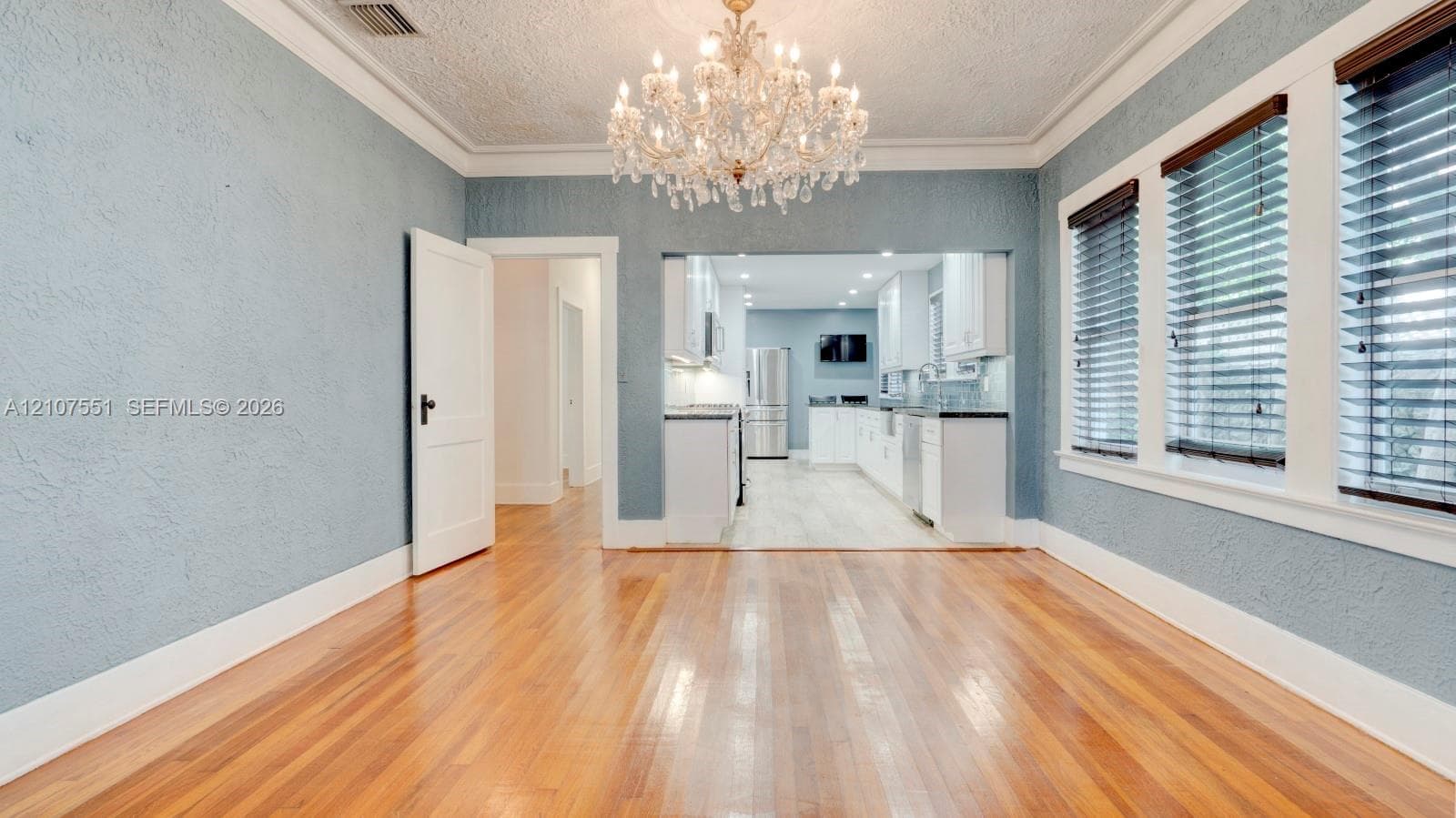 Hardwood flooring, ornate crystal chandelier, textured walls, crown molding, and multi-pane windows with horizontal blinds