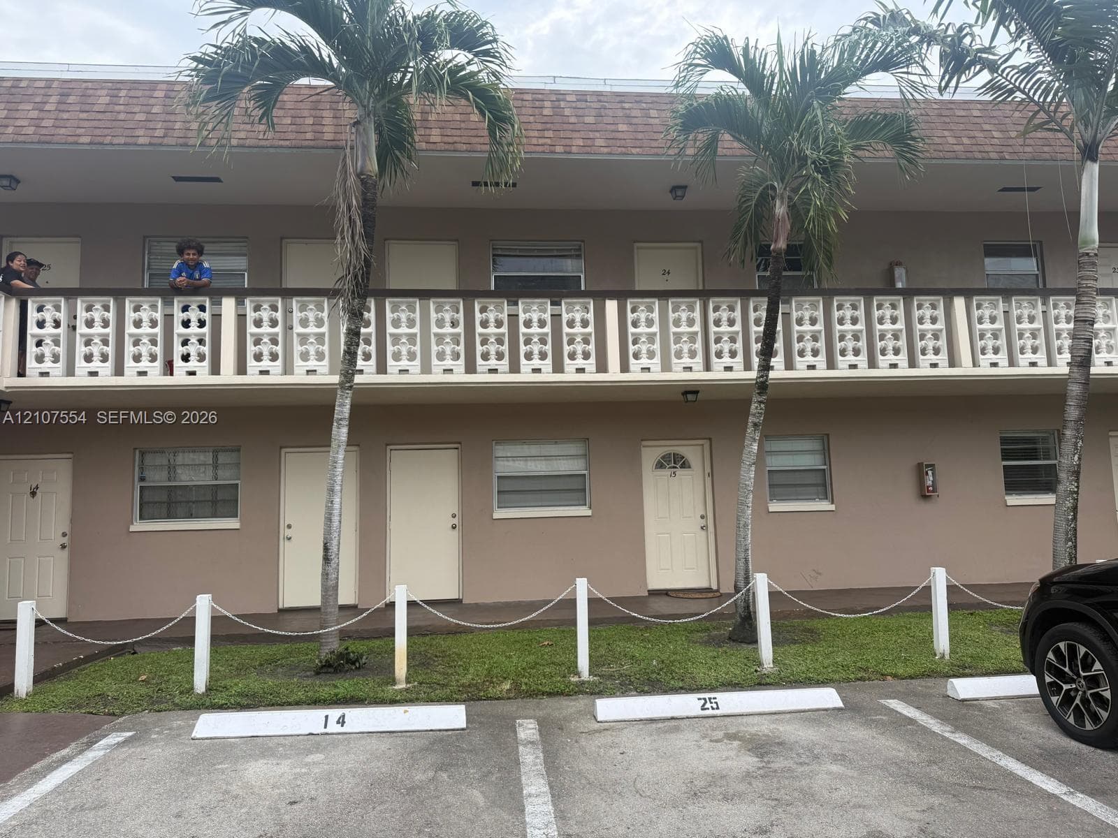 Two-story multi-unit exterior featuring decorative breeze block balcony railings, shingled mansard roofline, and assigned paved parking spaces